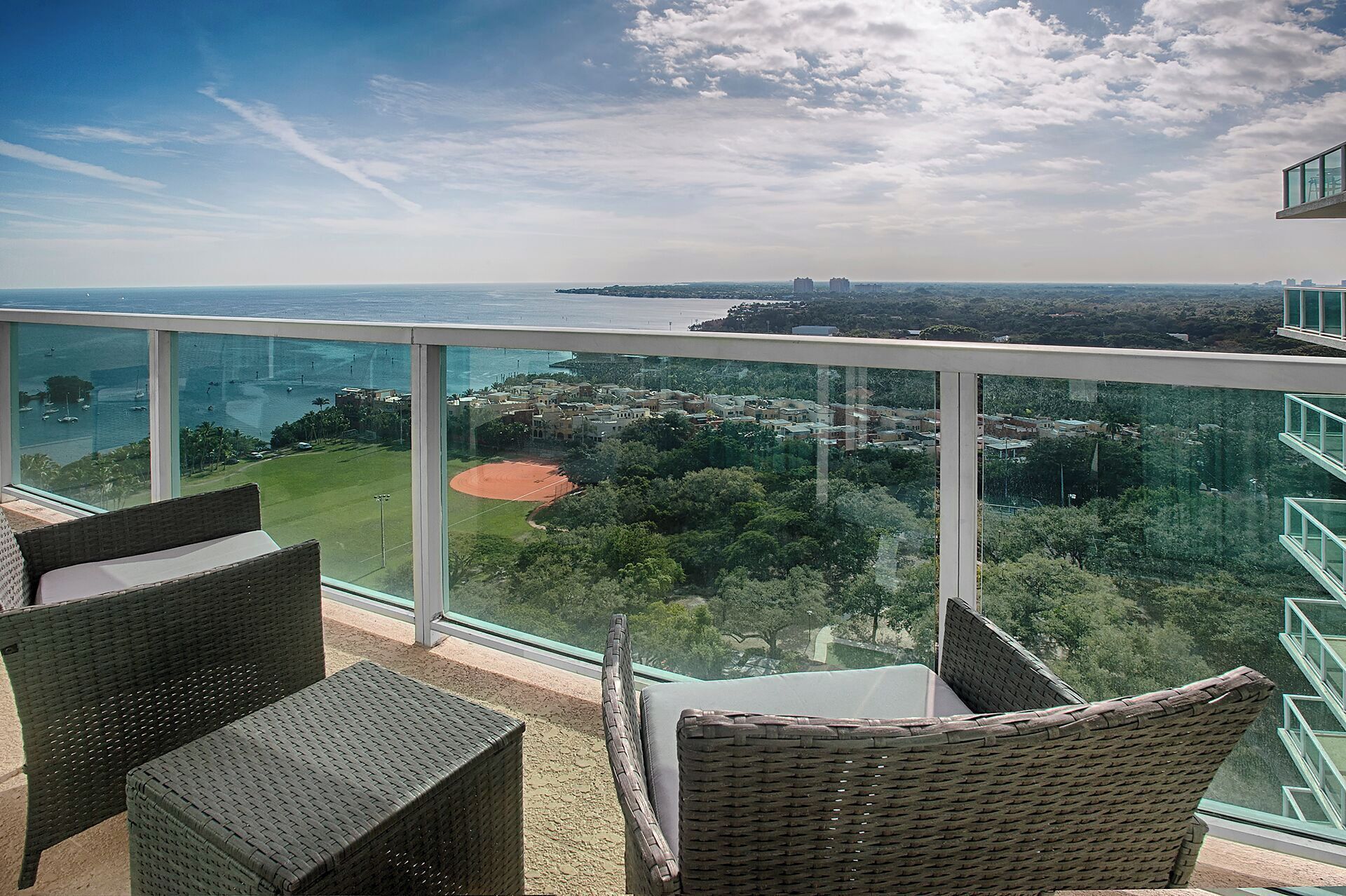 A balcony with wicker furniture overlooking a coastal view, including a baseball field and distant buildings.