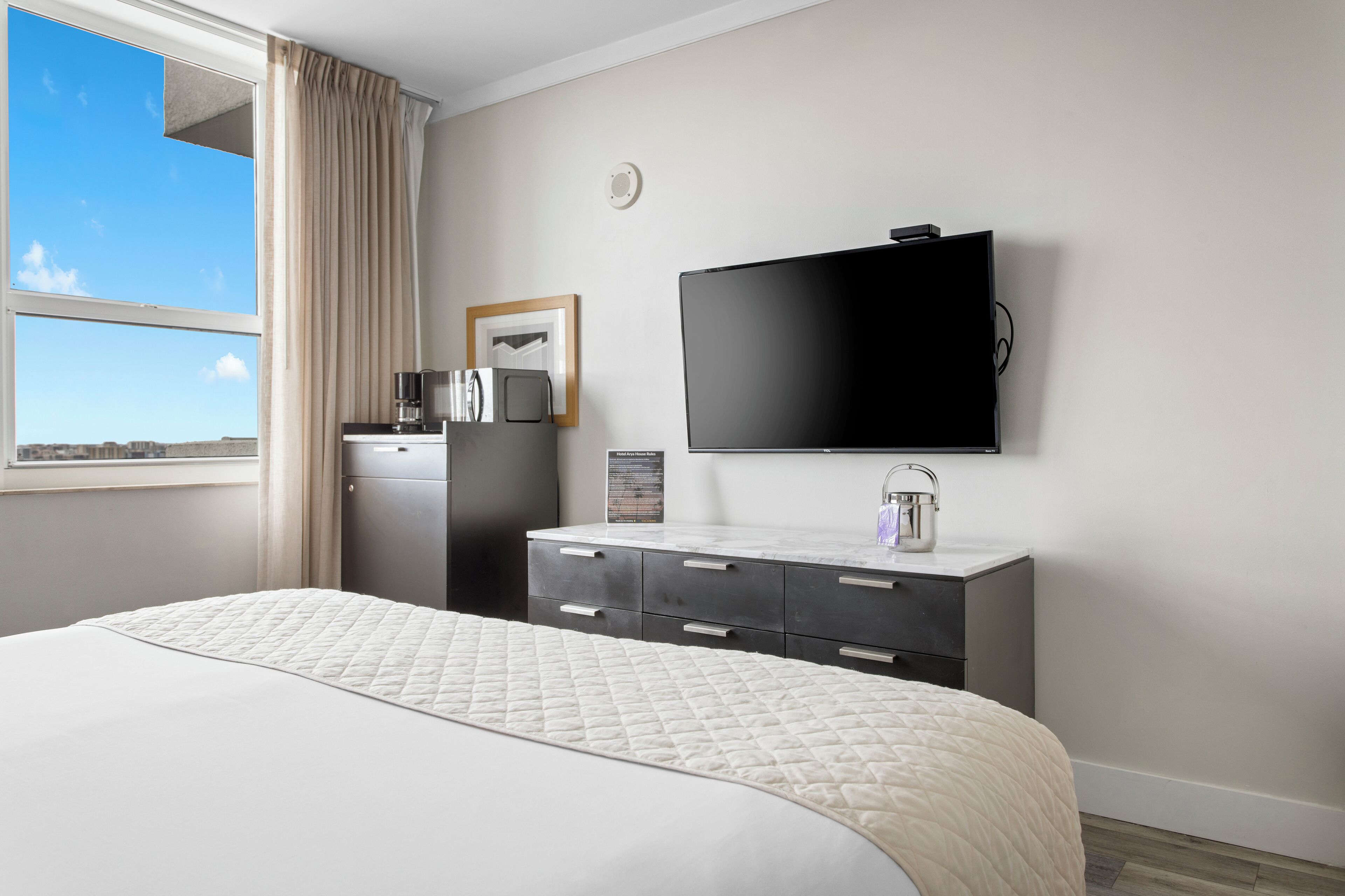 Quarto de hotel com uma cama grande, televisão de tela plana na parede, cómoda de cor escura, pequena mesa com cafeteira e janela com cortinas.