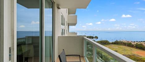 A balcony with a chair, glass doors, and a view of the sea and distant buildings.