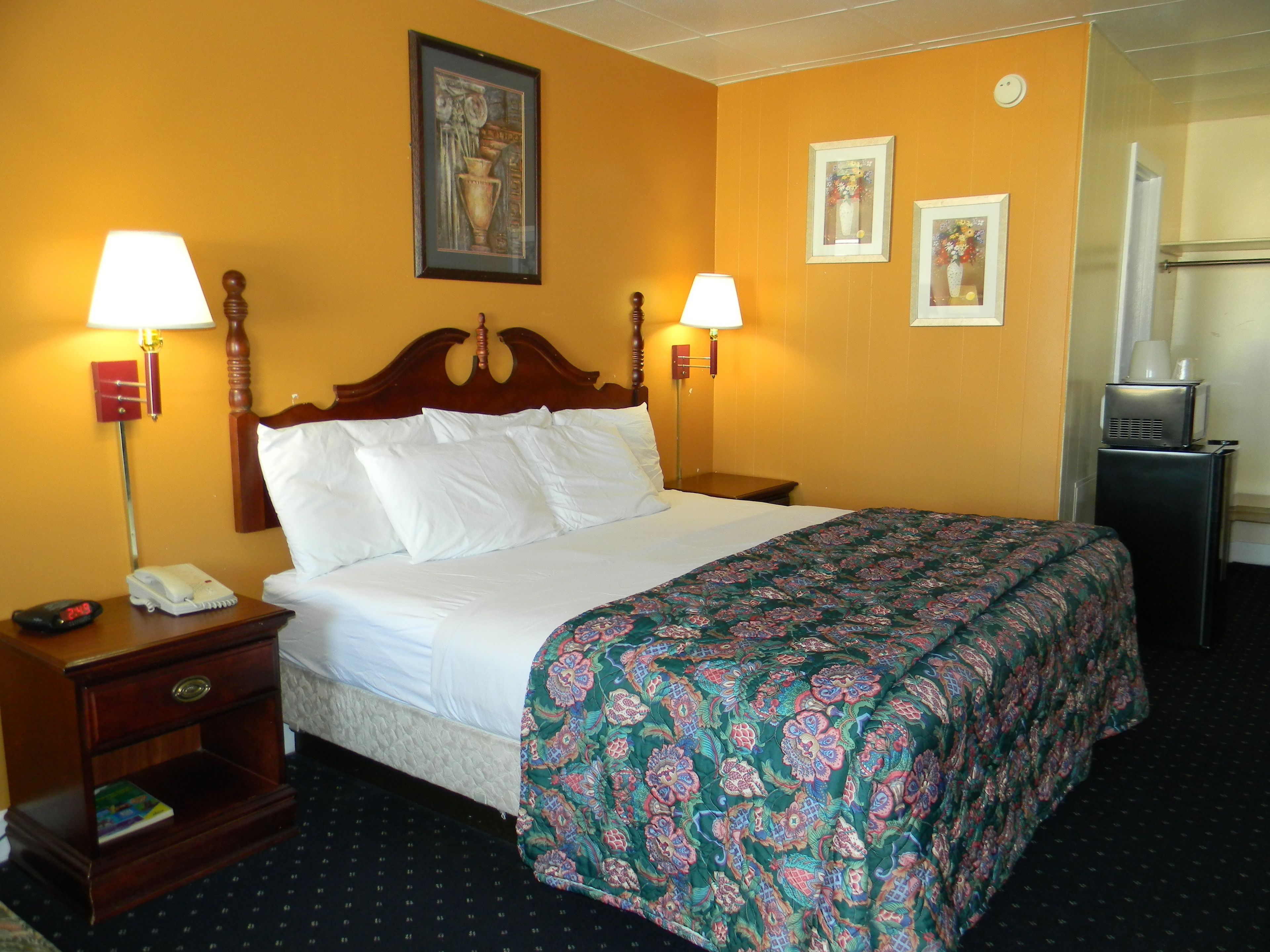 A hotel room with a bed, two bedside lamps, a nightstand with a phone, a small table, and a refrigerator.