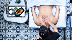 Eine Person erhält eine Rückenmassage auf einer Massageliege, in der Nähe stehen ein Obstkorb und ein Glas Milch.