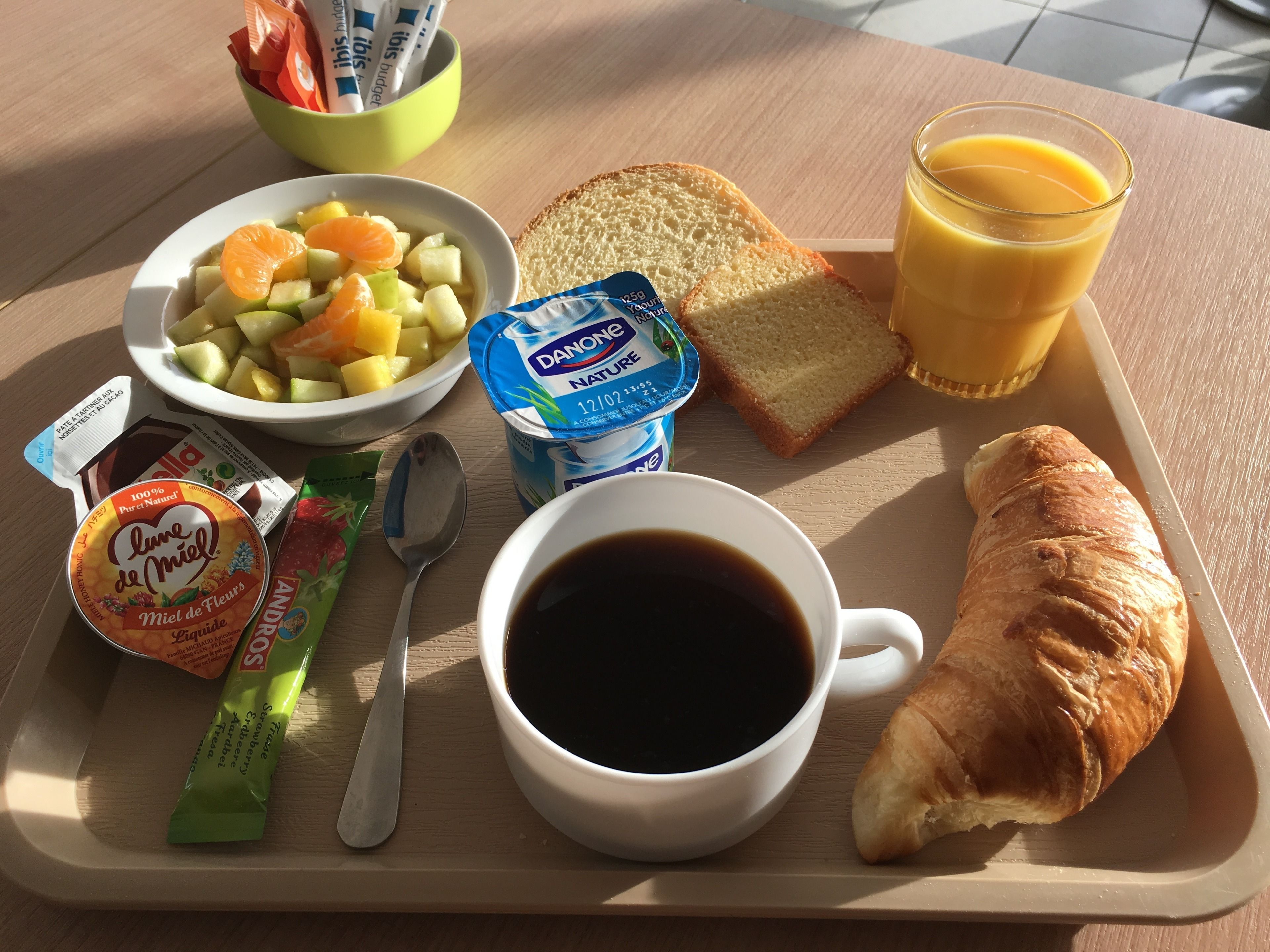 Un plateau de petit-déjeuner avec un bol de fruits, un verre de jus d’orange, un croissant, une tasse de café et un pot de yaourt Danone.