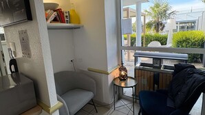 A small, modern room with a grey sofa, a small table with a lamp, a window with a view of the outside, and a shelf with books and a bottle. - ibis budget Ancenis (Mesanger)