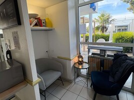 Quarto pequeno e moderno com um sofá cinzento, uma mesinha com abajur, uma janela com vista para o exterior e uma estante com livros e uma garrafa.