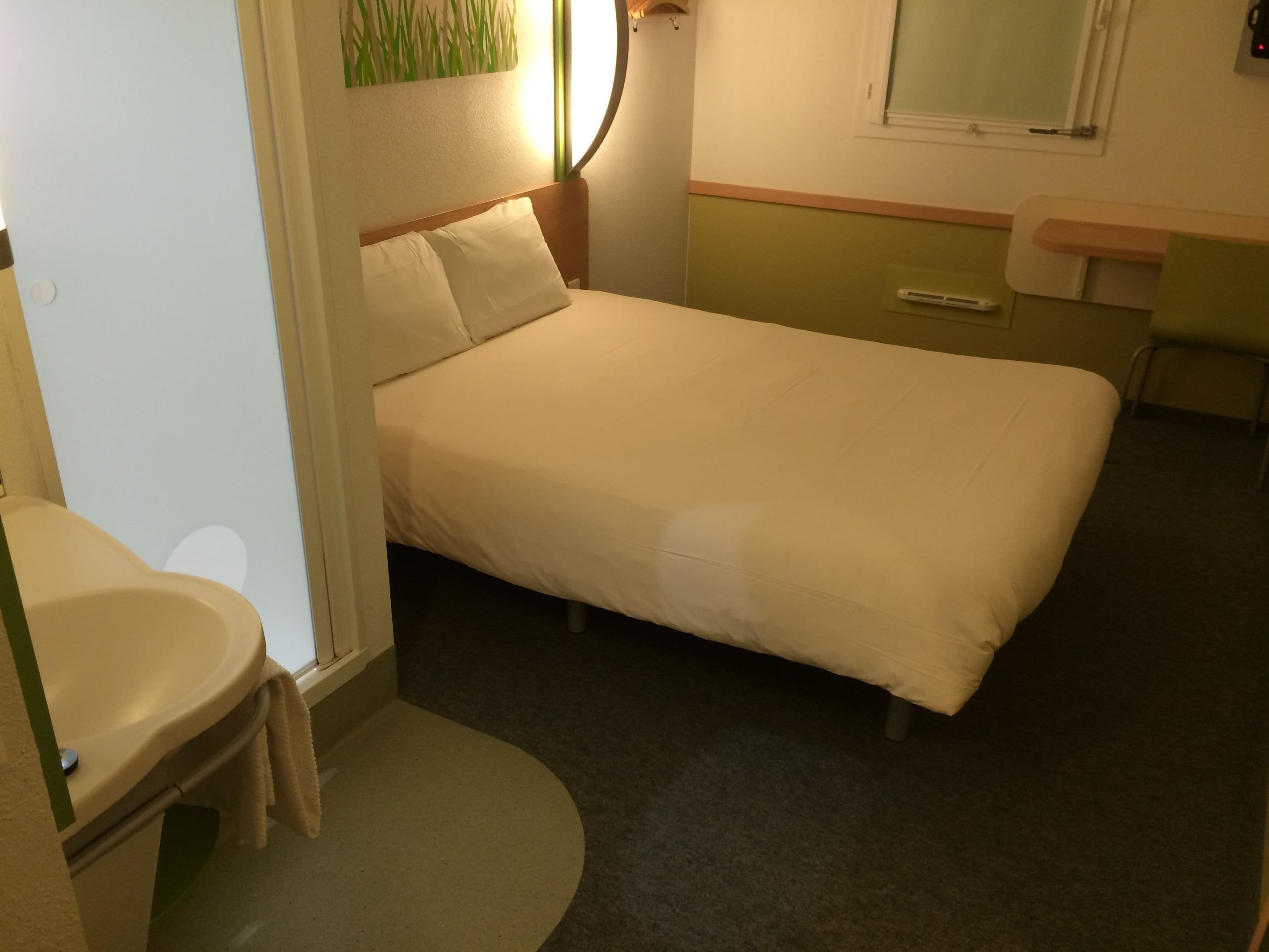 A small, single-bed room with a bedside table, a chair, a small desk, and a mirror on the wall.