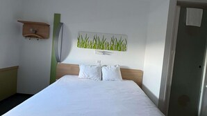 A hotel room with a bed, a wall-mounted artwork of green plants, a wooden headboard, and a door. - ibis budget Ancenis (Mesanger)