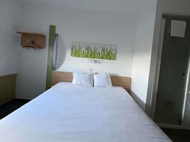 Quarto de hotel com cama, obra de arte na parede com plantas verdes, cabeceira de madeira e uma porta.
