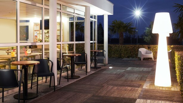 An outdoor patio area with tables and chairs, a large illuminated lamp, and a building with large windows. - ibis budget Ancenis (Mesanger)