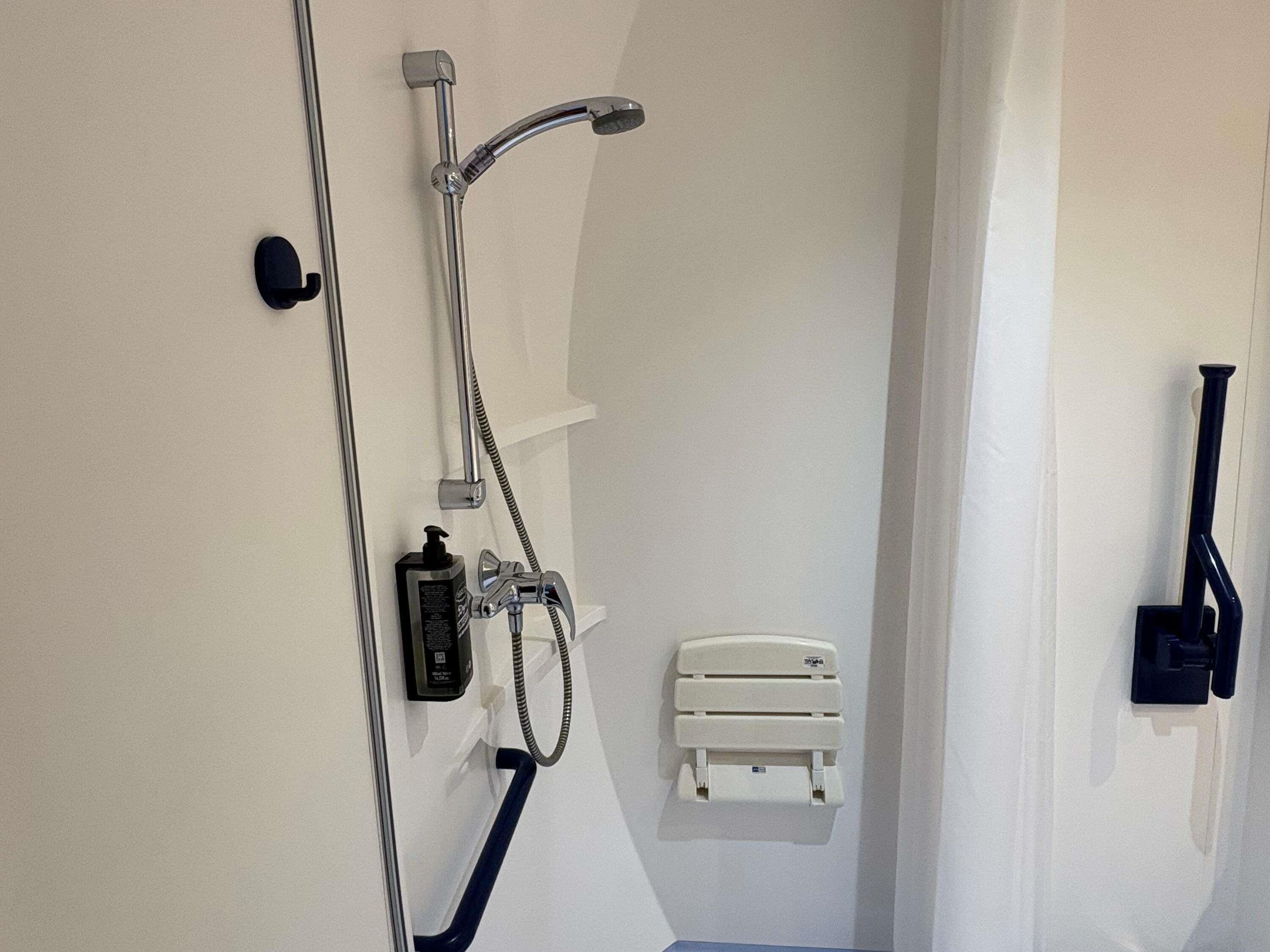Une salle de bain avec une douche, un distributeur de savon, une petite étagère blanche et un aspirateur fixé au mur.