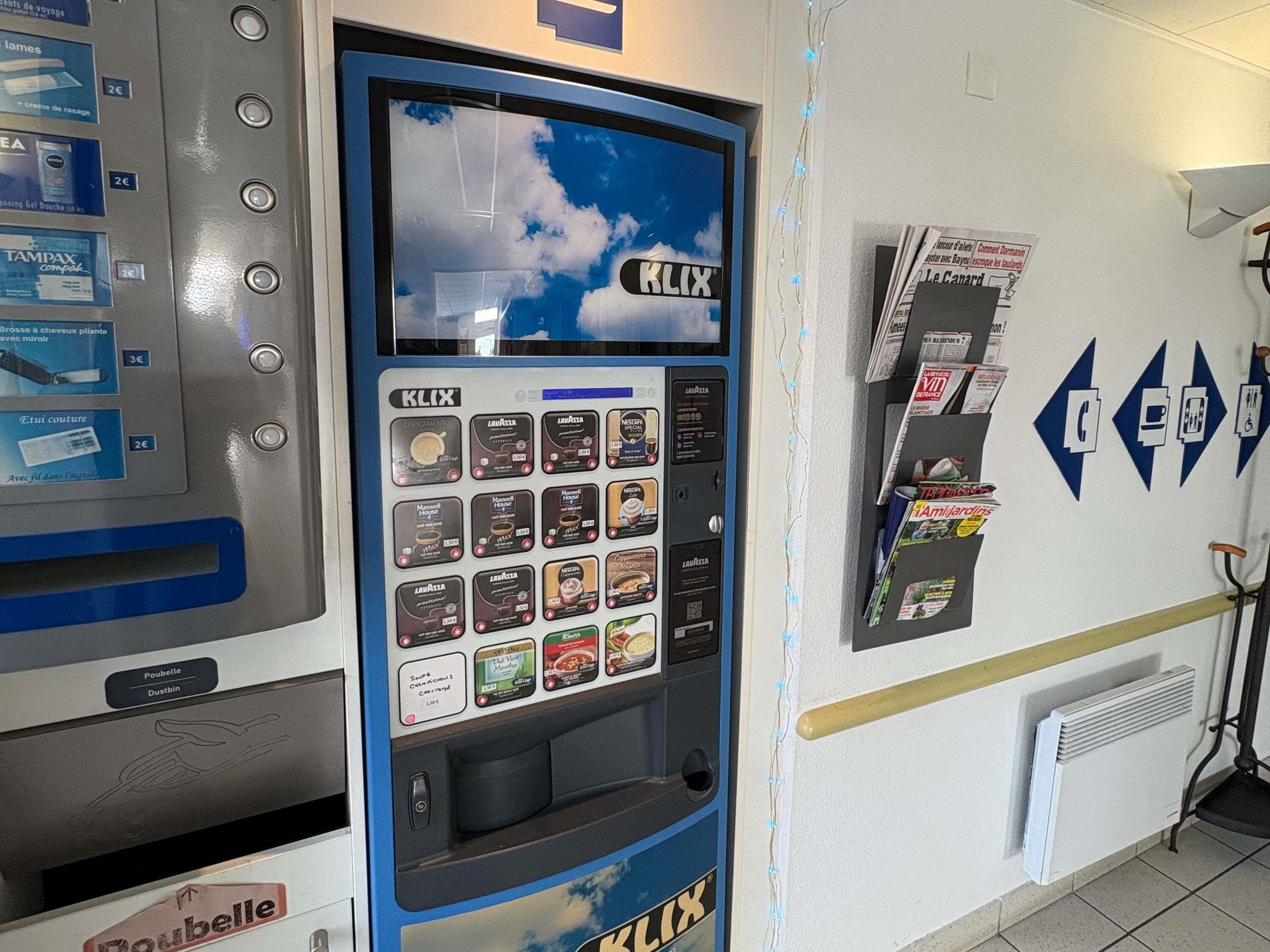 En automattankstation med en skärm som visar en himmel och moln, omgiven av olika sorters drycker.
