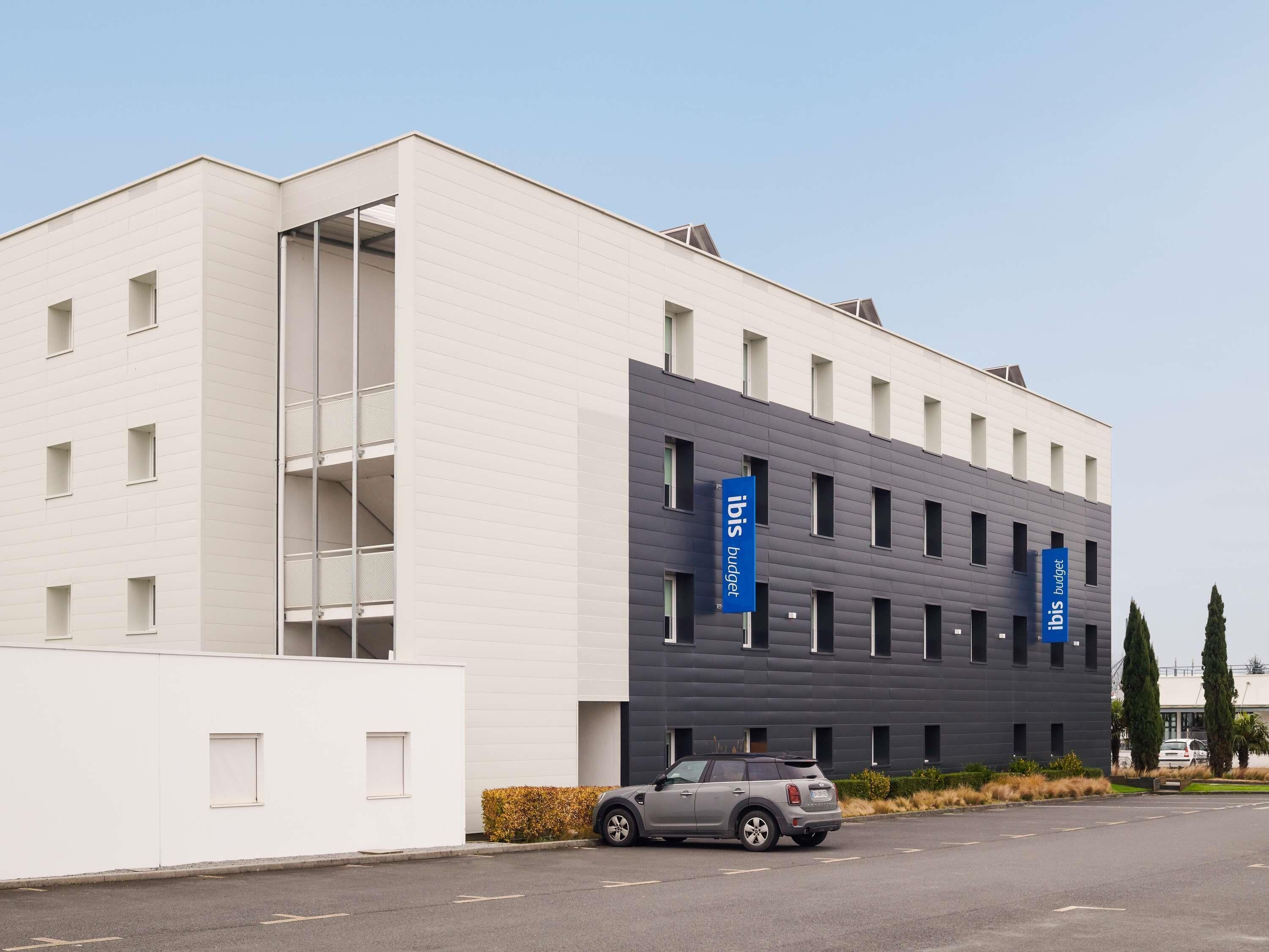 Un edificio moderno con fachada blanca y sección gris, con dos banderines azules que muestran el texto 'ibis budget'.