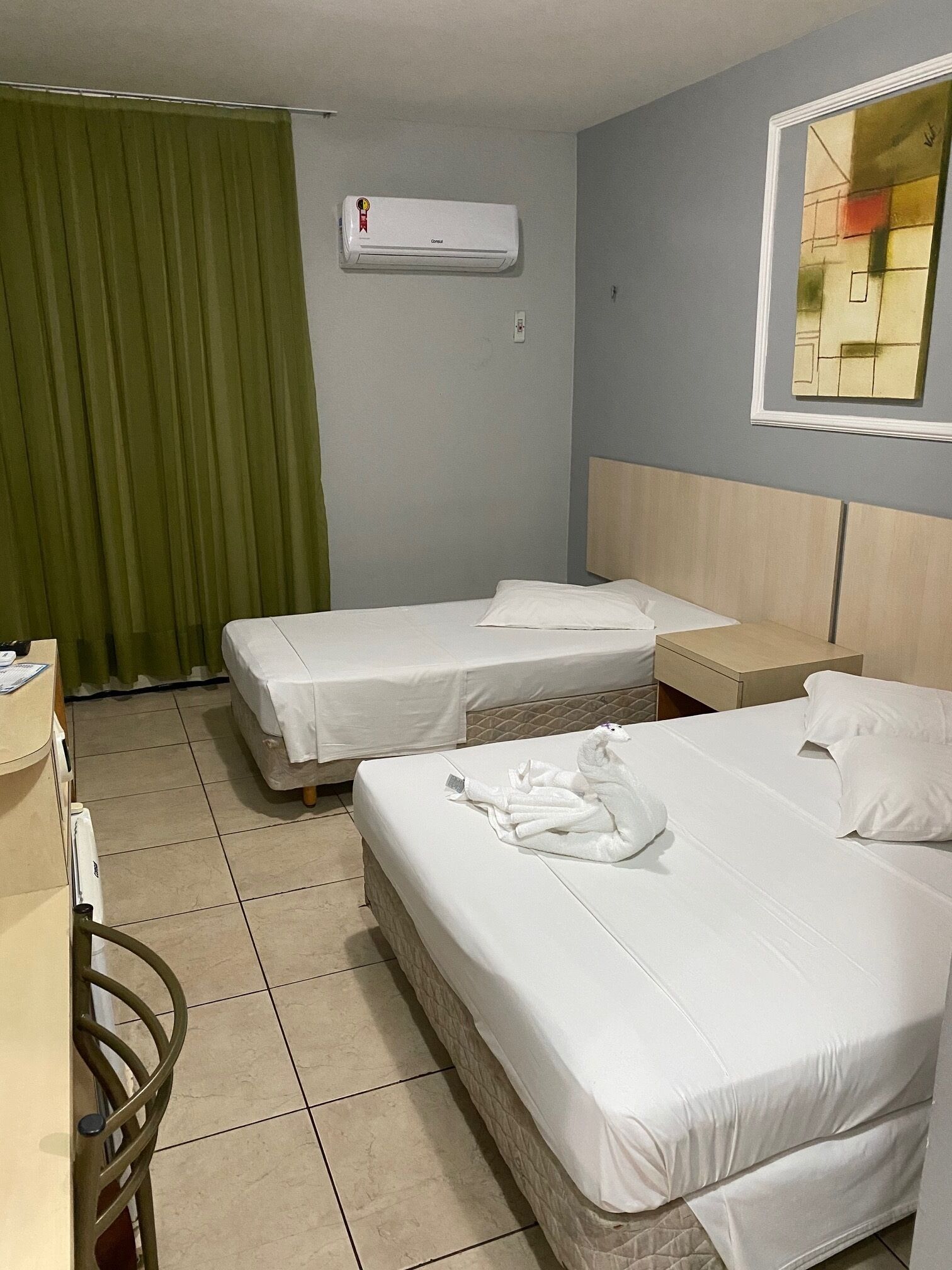 A hotel room with two beds, a green curtain, an air conditioning unit, a wooden headboard, and a small table.