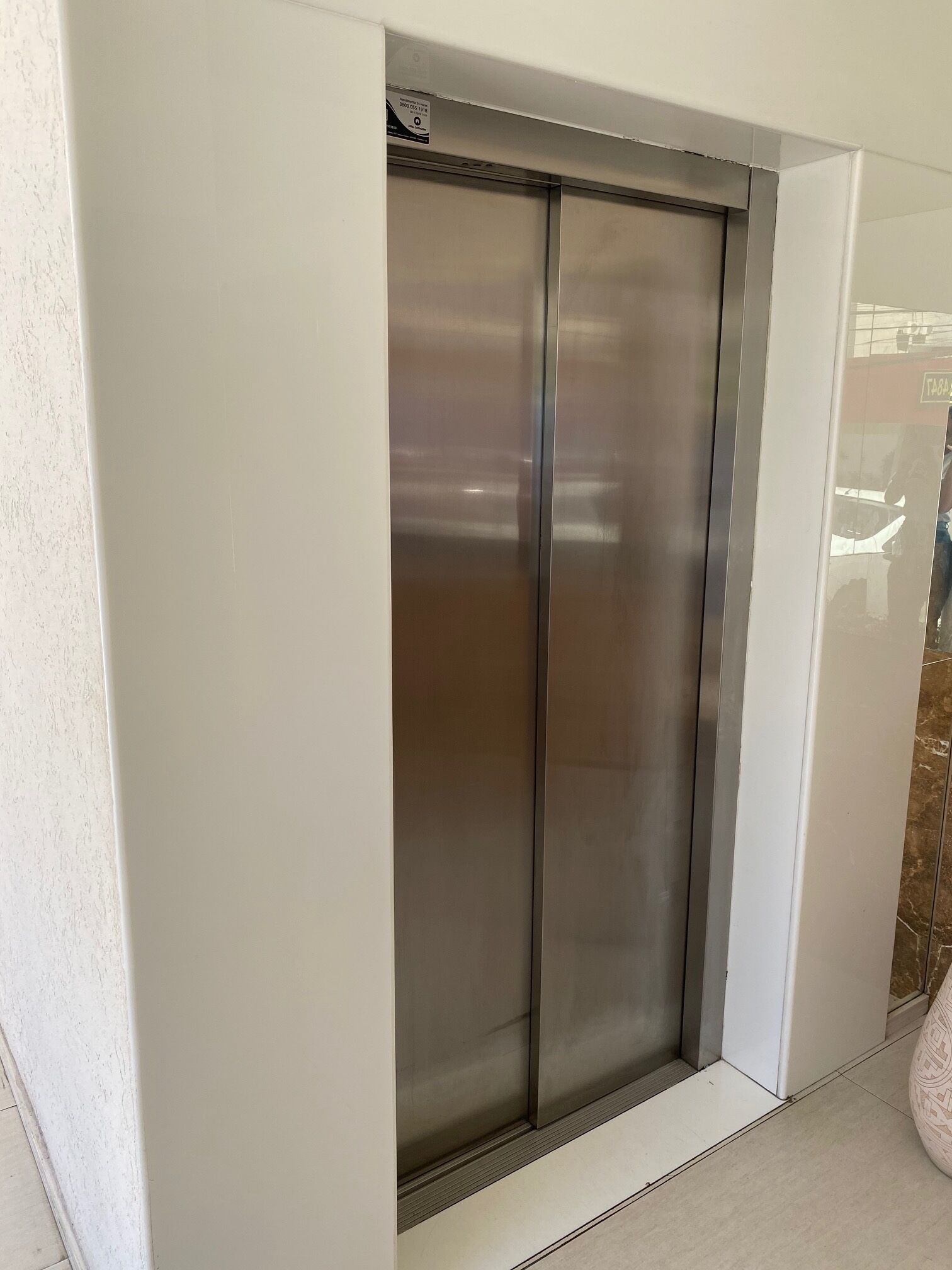 A modern elevator with a metallic finish, located in a hallway with white walls and a tiled floor.