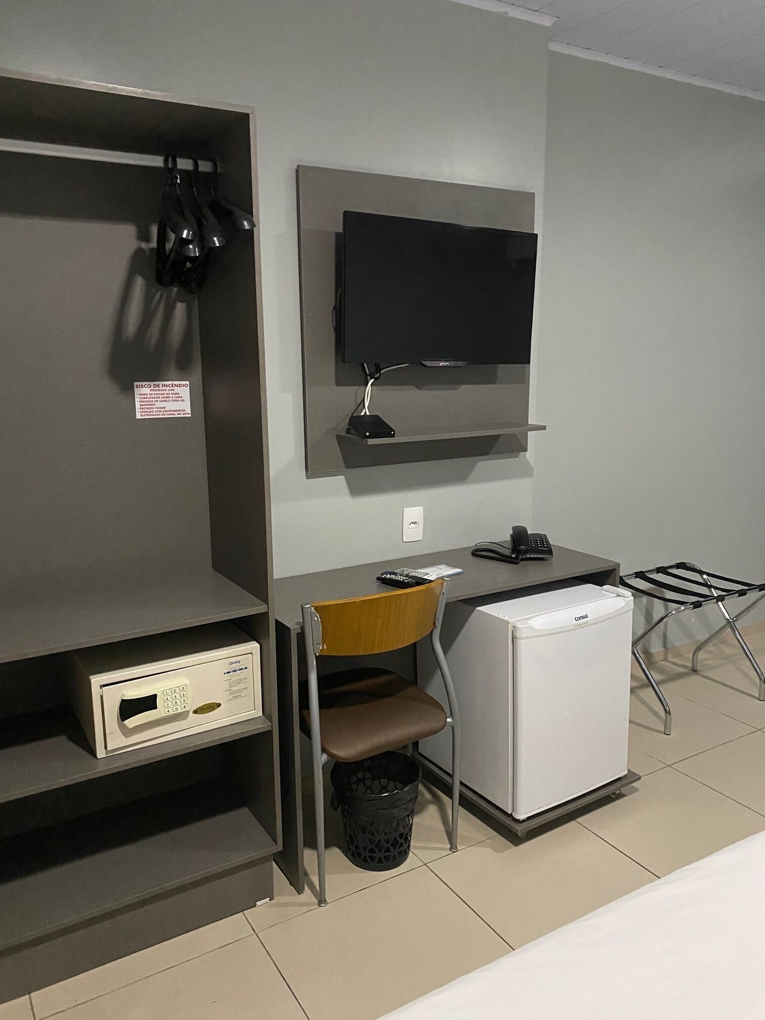 A compact hotel room with a kitchenette, a small refrigerator, a desk with a chair, a television mounted on the wall, and a shoe rack.