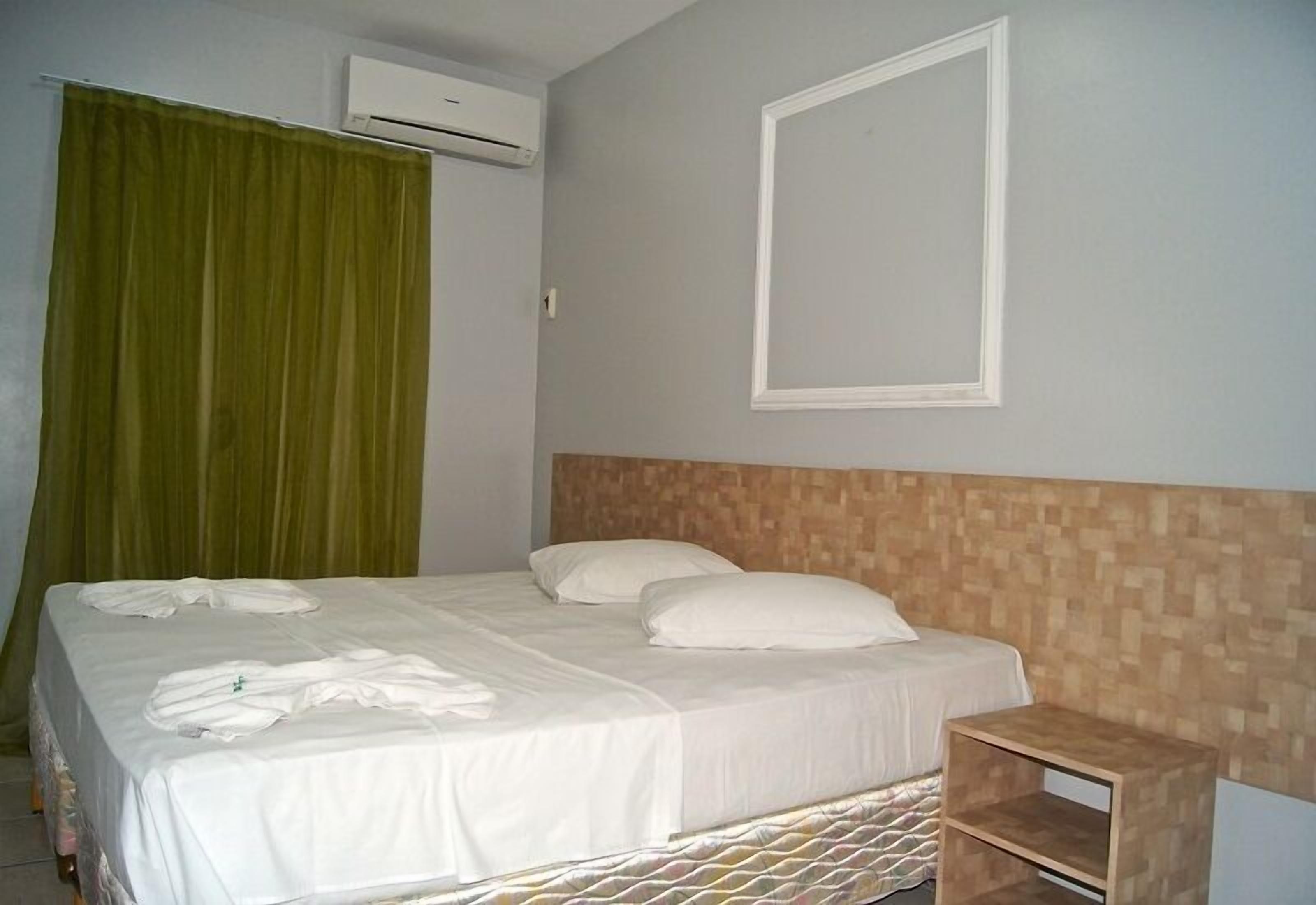 A neatly made bed with white linens and pillows, a wooden headboard, a small wooden nightstand, and a green curtain.