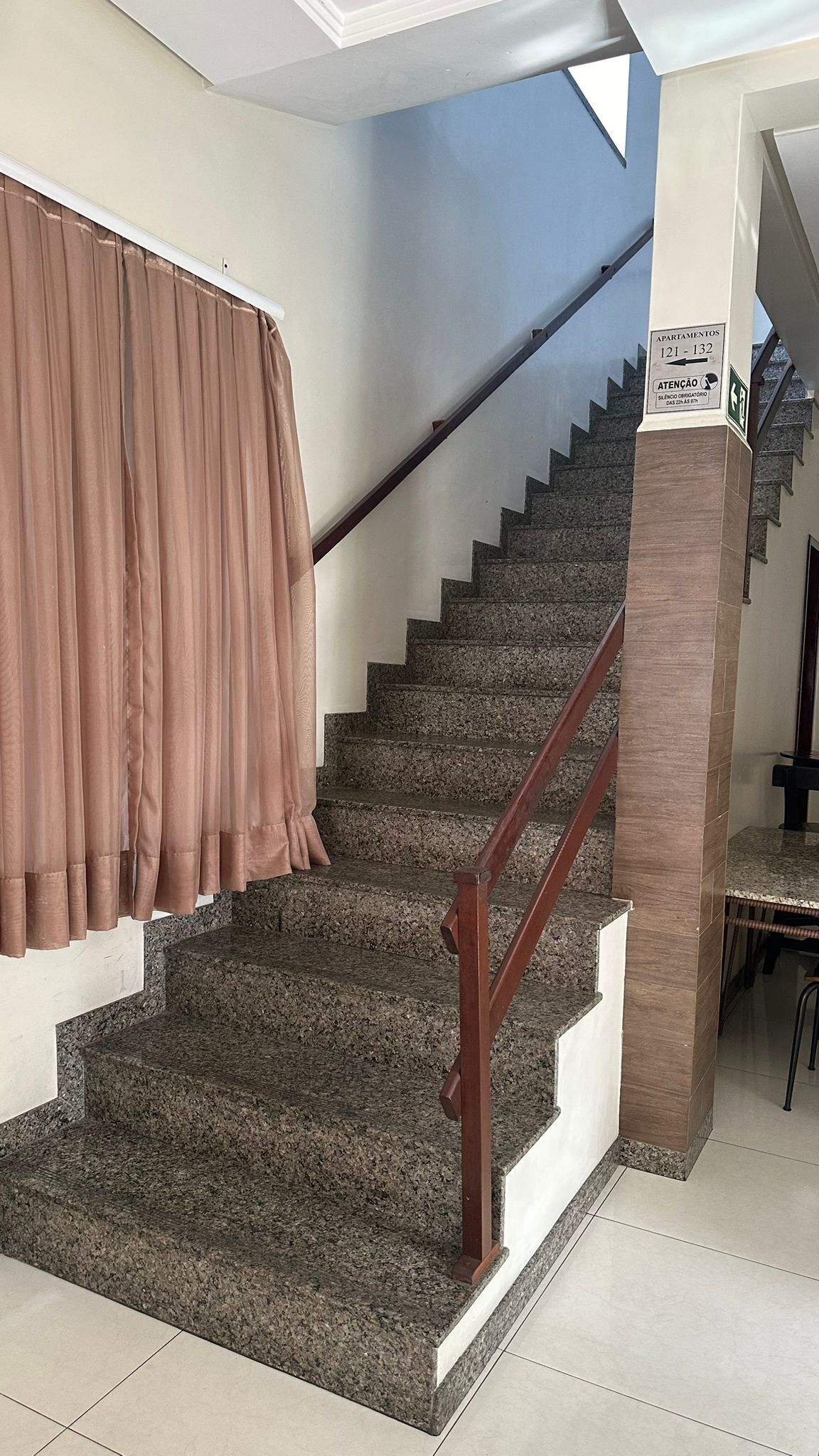 Uma escadaria com corrimão de madeira, degraus carpetados e cortina marrom.