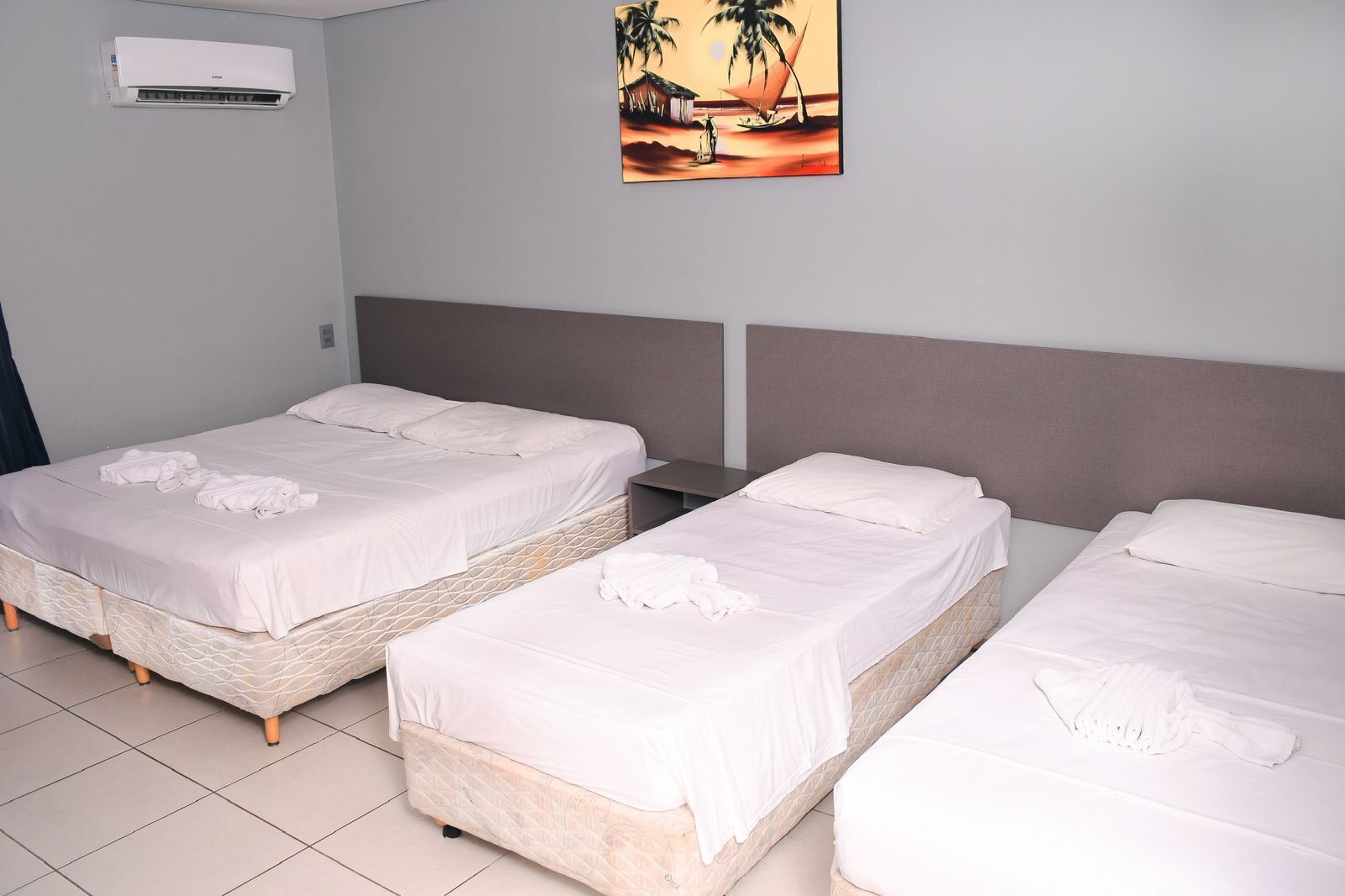 A hotel room with three beds, a painting of a sunset, and an air conditioning unit.