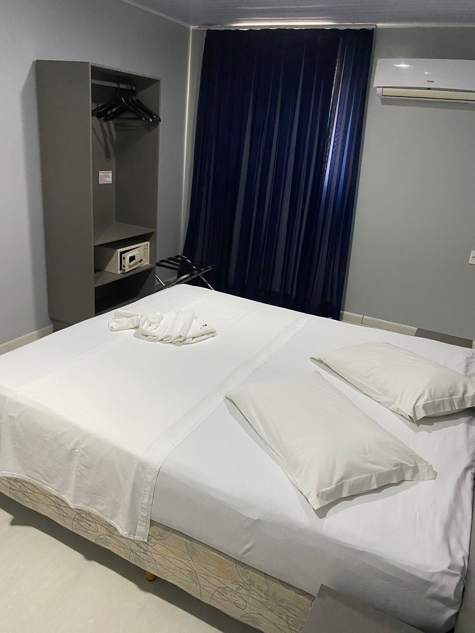 Un letto rifatto con lenzuola bianche, un armadio grigio con appendiabiti e una finestra con tende blu.