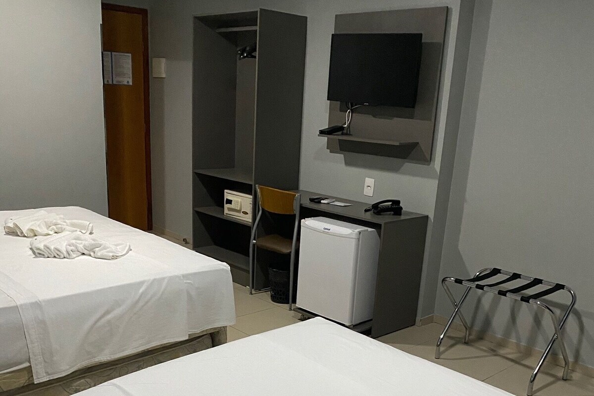 A hotel room with two beds, a desk, a chair, a small refrigerator, a TV mounted on the wall, and a coat rack.