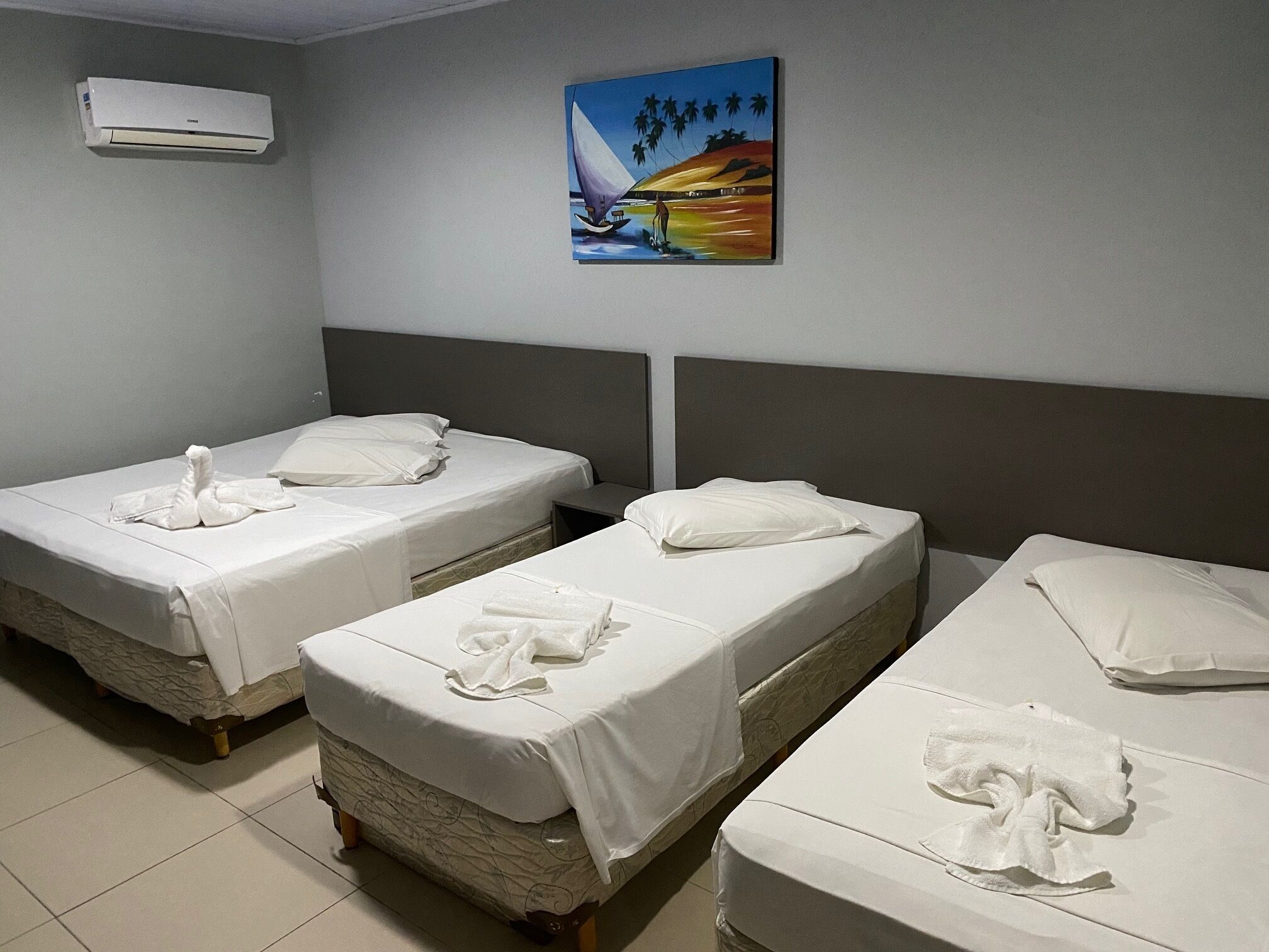 Habitación de hotel con tres camas individuales, cada una con una colcha blanca y una toalla. En la pared, sobre las camas, hay un cuadro de una escena tropical.