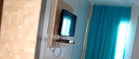 A hotel room with a tiled floor, a desk with a telephone, a television mounted on the wall, and a blue curtain.