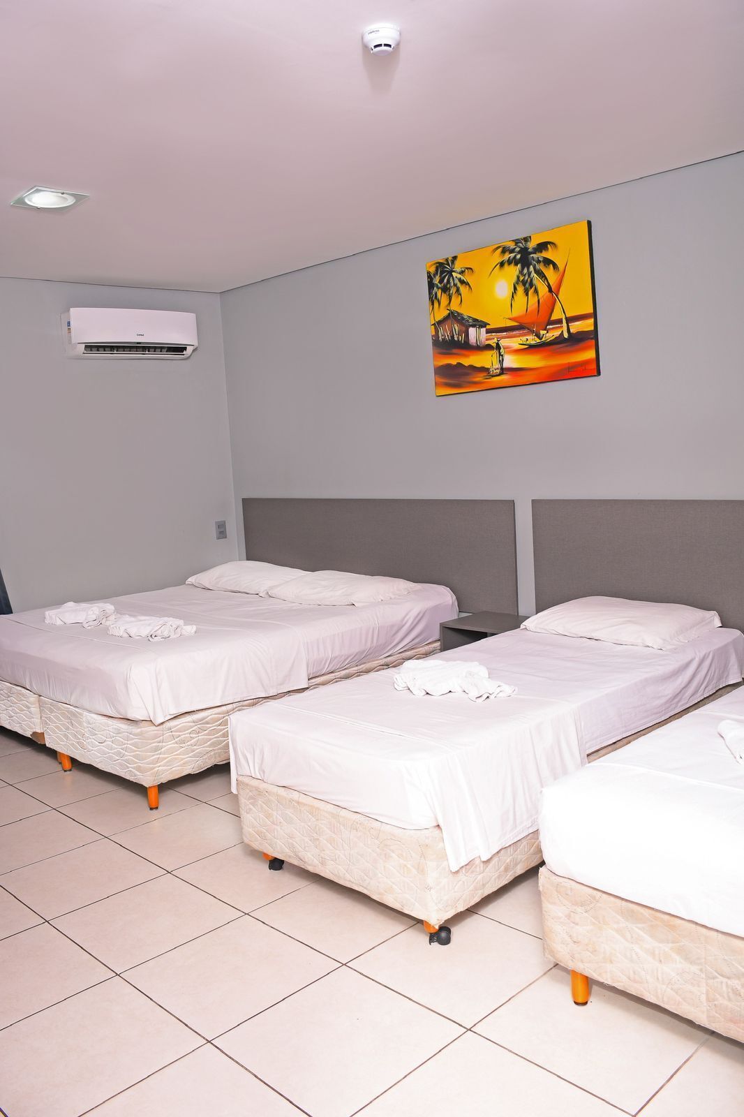 A hotel room with three beds, a ceiling-mounted air conditioner, and a painting of a sunset scene.