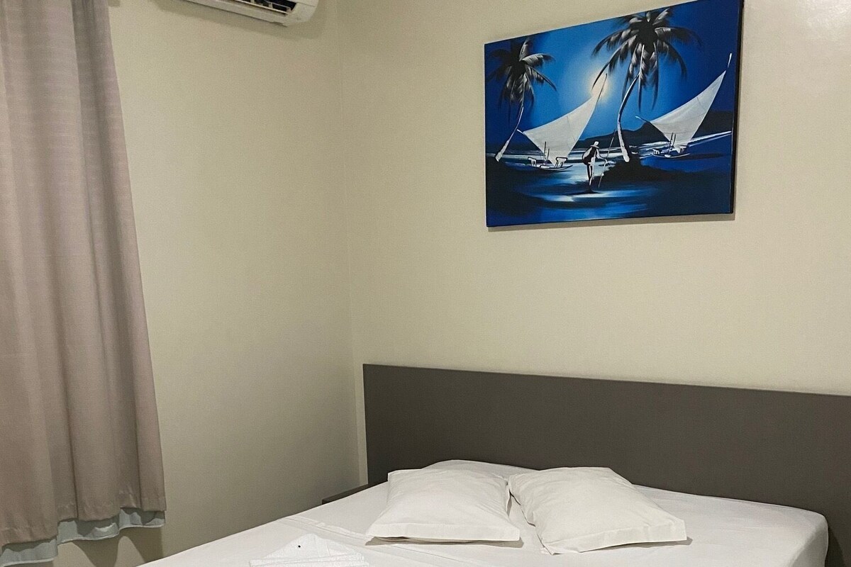 Una cama bien hecha con sábanas blancas, un cuadro de un velero en la pared y un equipo de aire acondicionado sobre la cama.