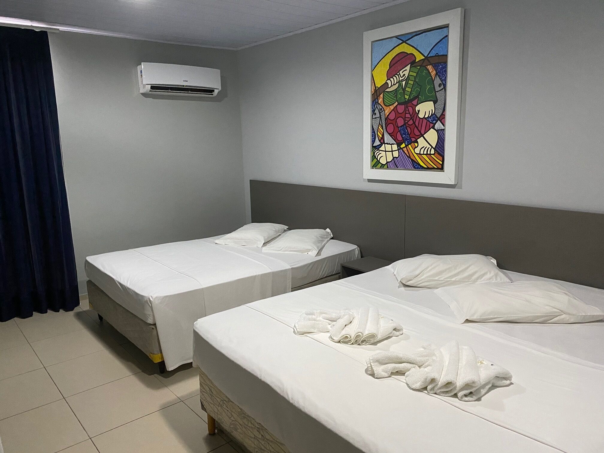Habitación de hotel con dos camas individuales, un ventanal con cortinas, aire acondicionado montado en la pared y una obra de arte colorida sobre las camas.