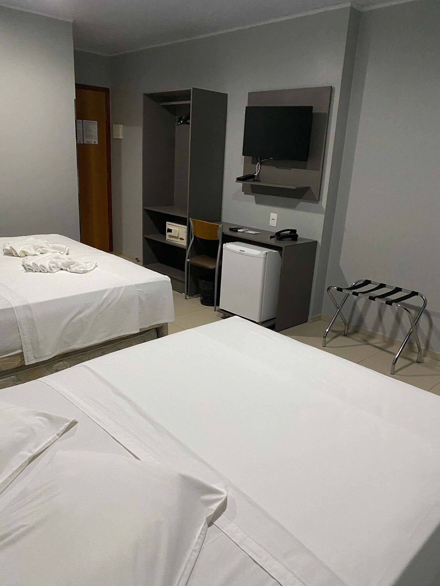 A hotel room with two beds, a desk, a chair, a small refrigerator, and a TV mounted on the wall.