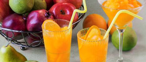 Uma fruteira com maçãs, laranjas e limões, e dois copos de suco de laranja com canudos.