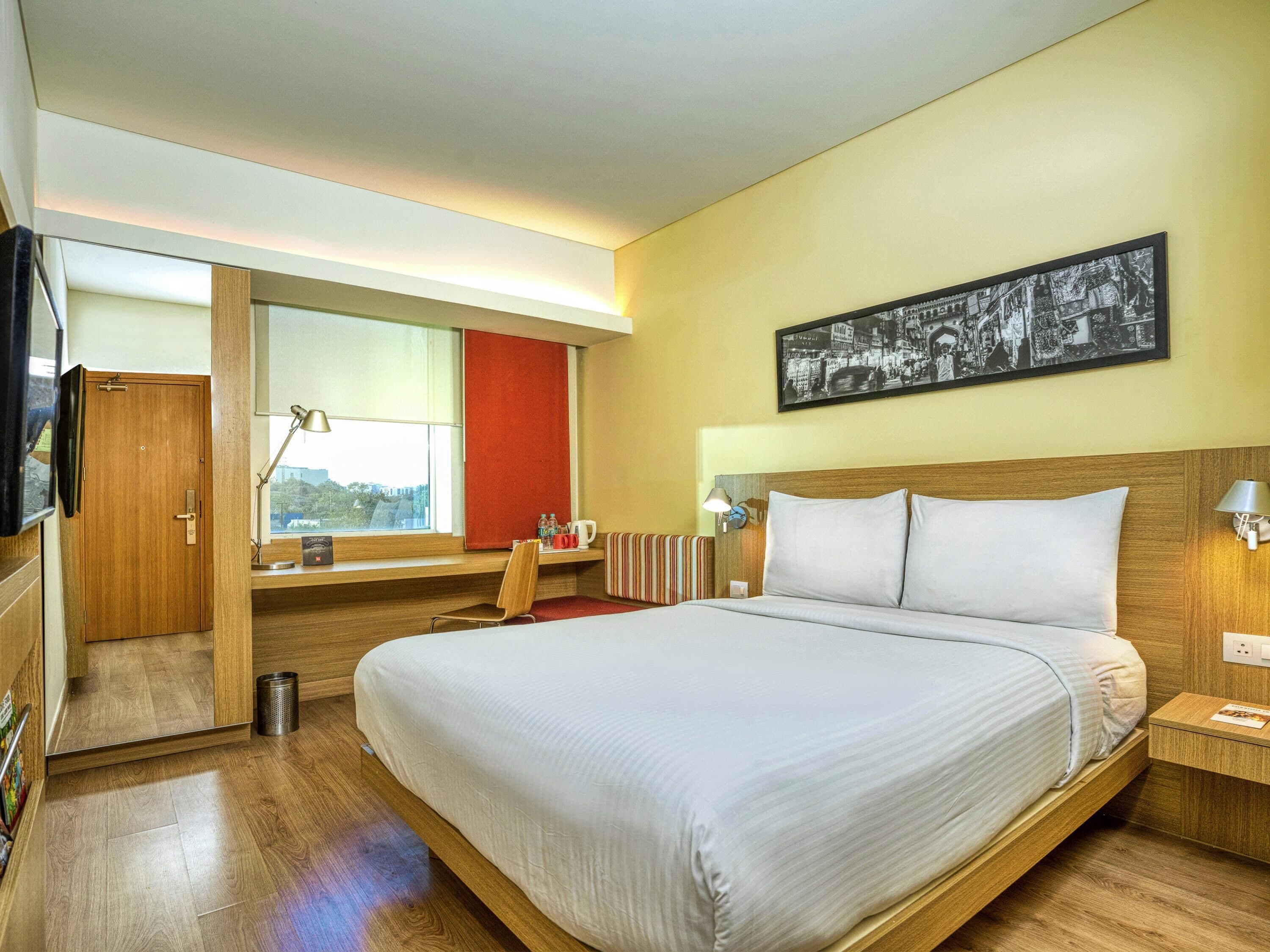 Quarto de hotel com uma cama grande, uma secretária com cadeira, janela com vista, guarda-roupa de madeira e televisão na parede.