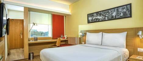 Quarto de hotel com uma cama grande, uma secretária com cadeira, janela com vista, guarda-roupa de madeira e televisão na parede.