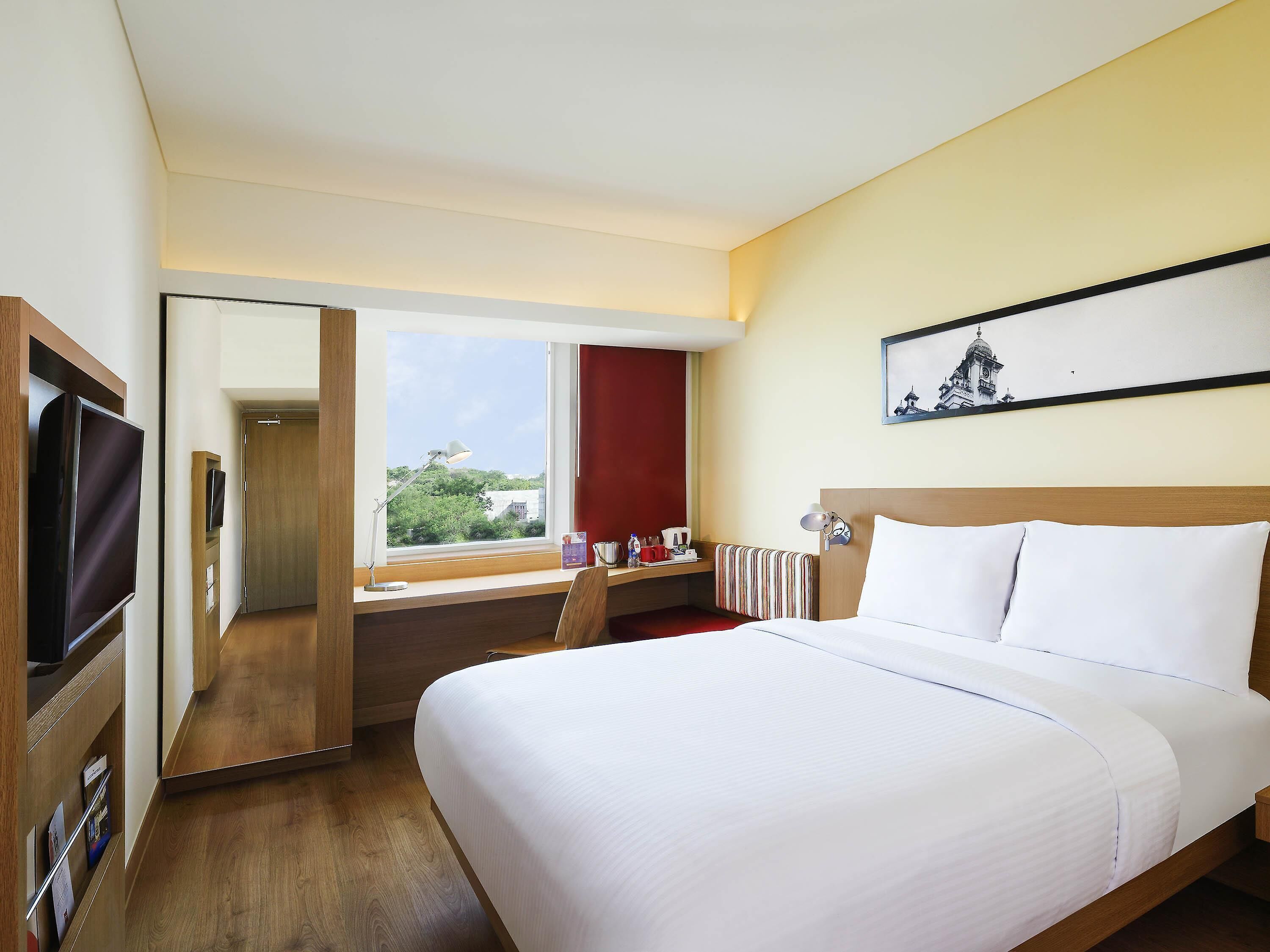 A hotel room with a bed, a desk with a computer, a TV, a window with a view, and a painting on the wall.