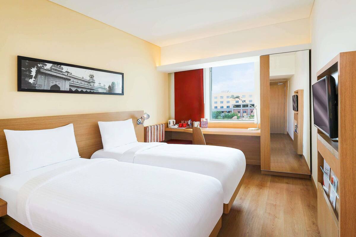 Habitación de hotel con dos camas, televisión, escritorio, ventana con vistas y un cuadro en la pared.