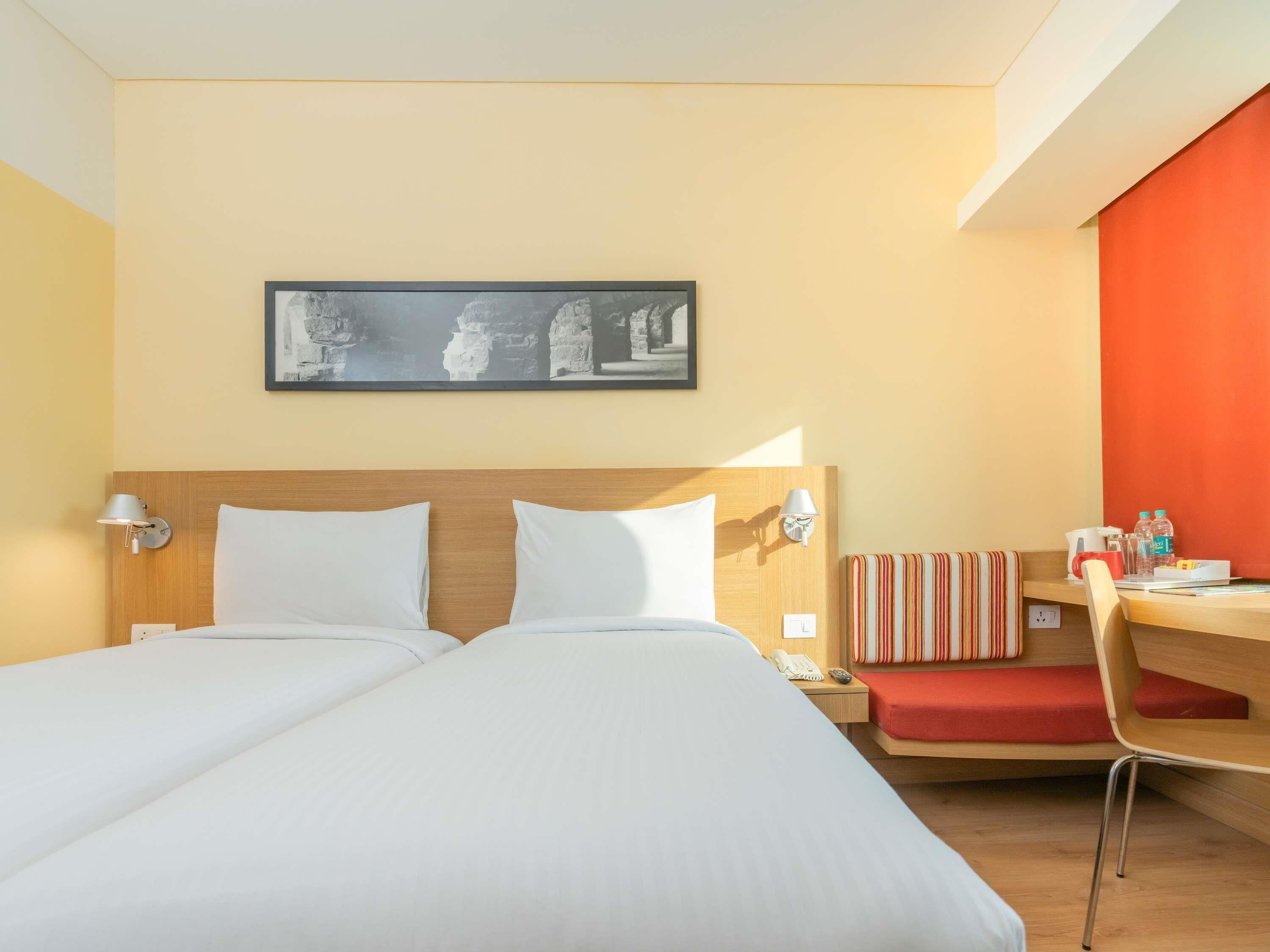 A hotel room with two beds, a desk with a chair, a painting on the wall, and a red accent wall.