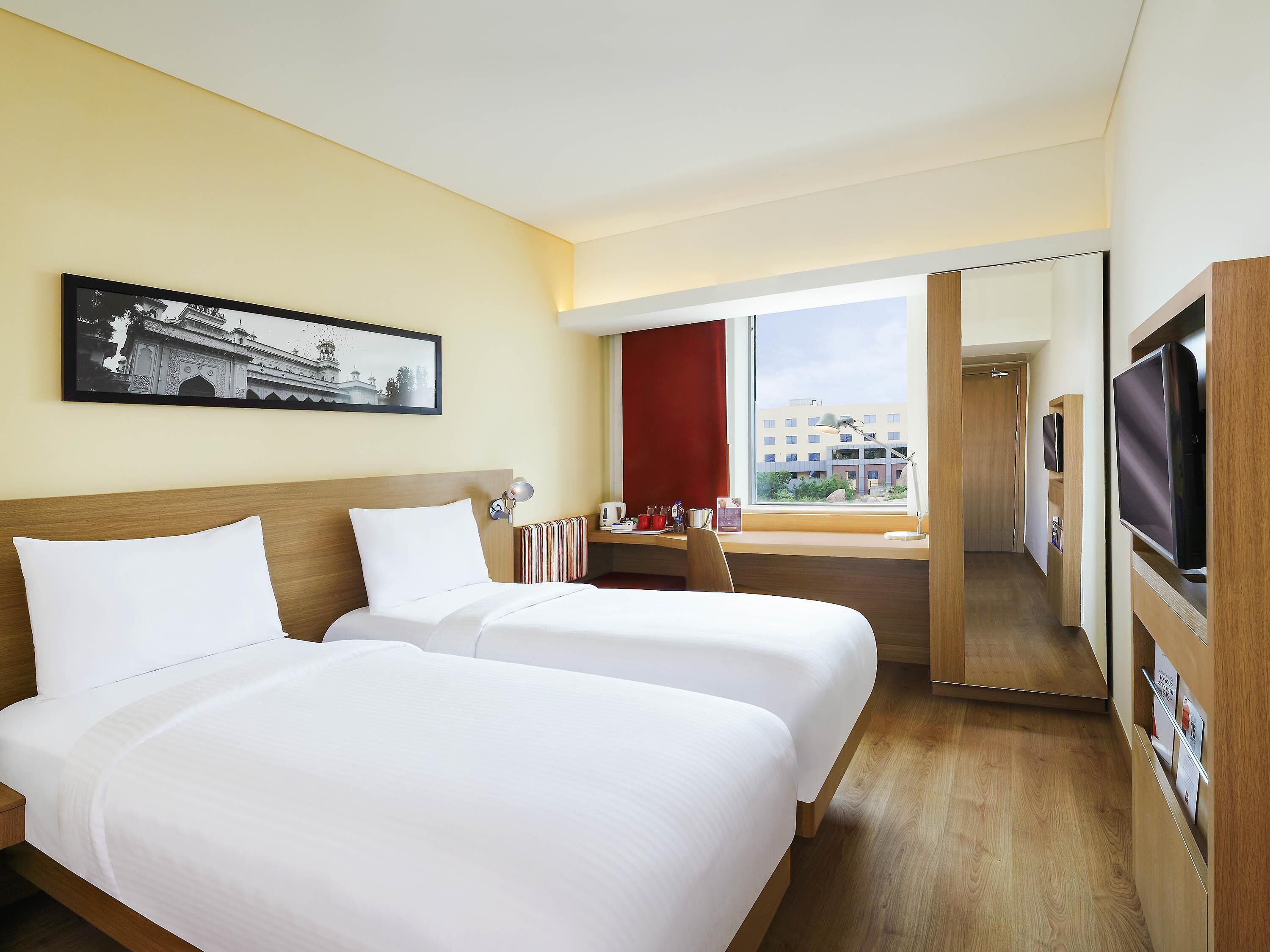 A hotel room with two beds, a TV, a desk, a window with a view of buildings, and a framed picture on the wall.
