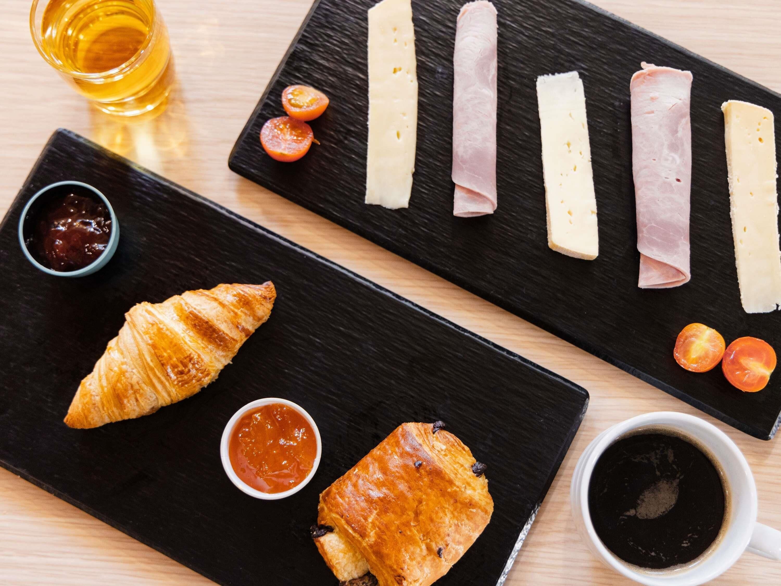 Eine schwarze Schiefertafel mit einem Croissant, zwei kleinen Schalen Marmelade, einer Tasse Kaffee und einer Auswahl an Käse und Wurst.