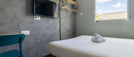 A modern hotel room with a bed, a television mounted on the wall, a window with a view of the sky, and a chair.