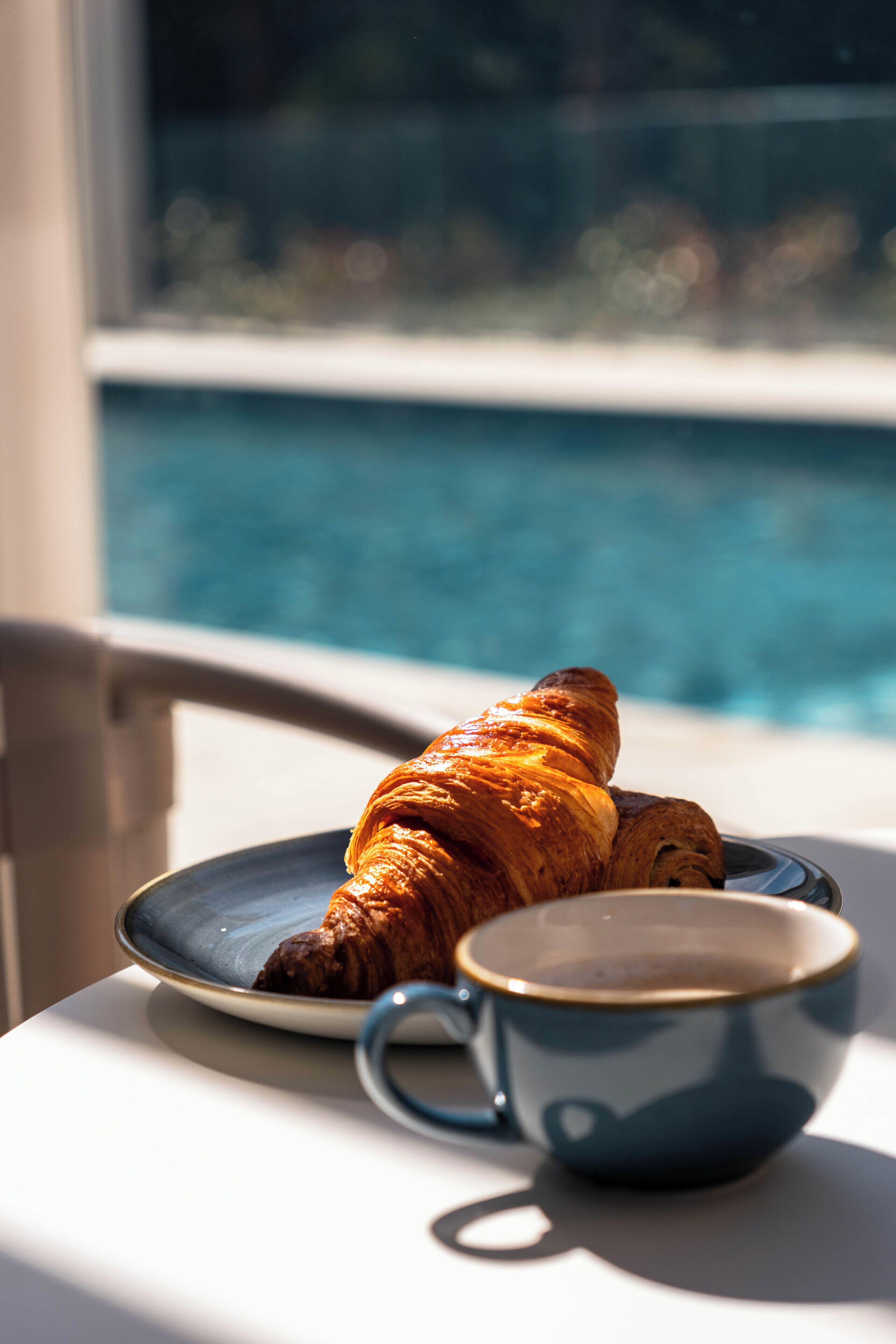 A croissant on a plate next to a cup, with a pool in the background.