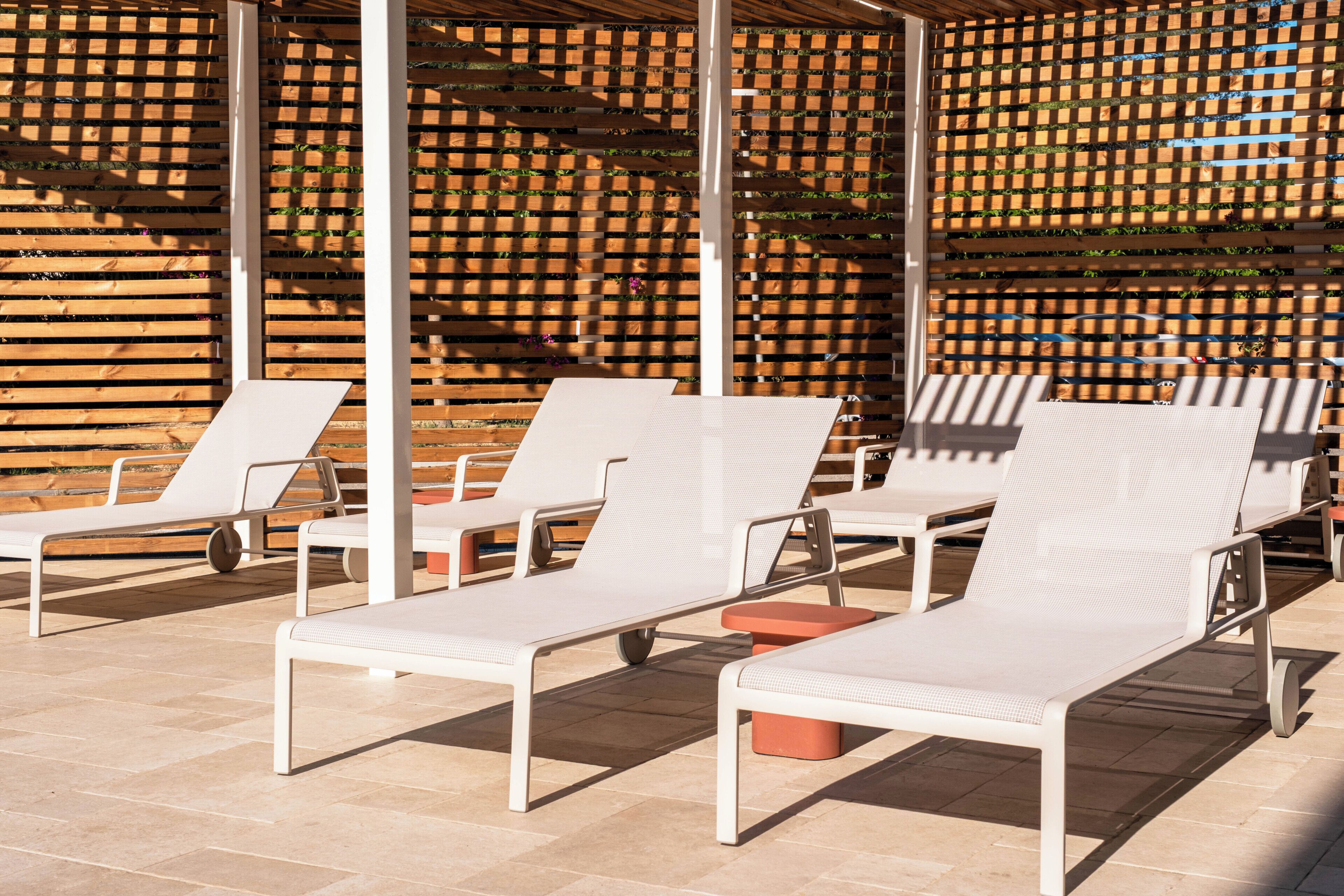 A sunlit outdoor seating area with white lounge chairs and a wooden lattice wall in the background.