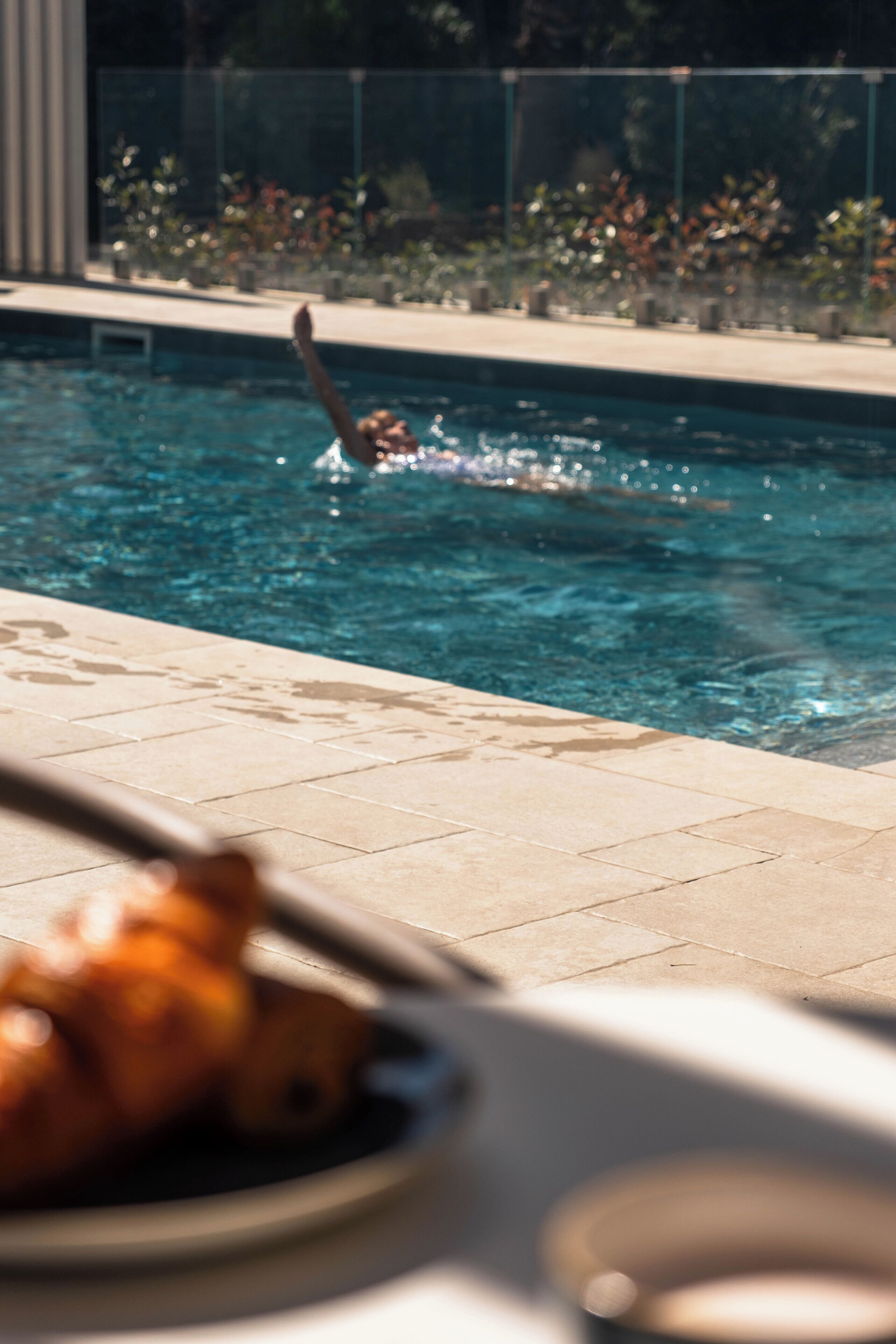 A swimming pool with a person diving, a tiled deck, and a dining table with a plate of food.