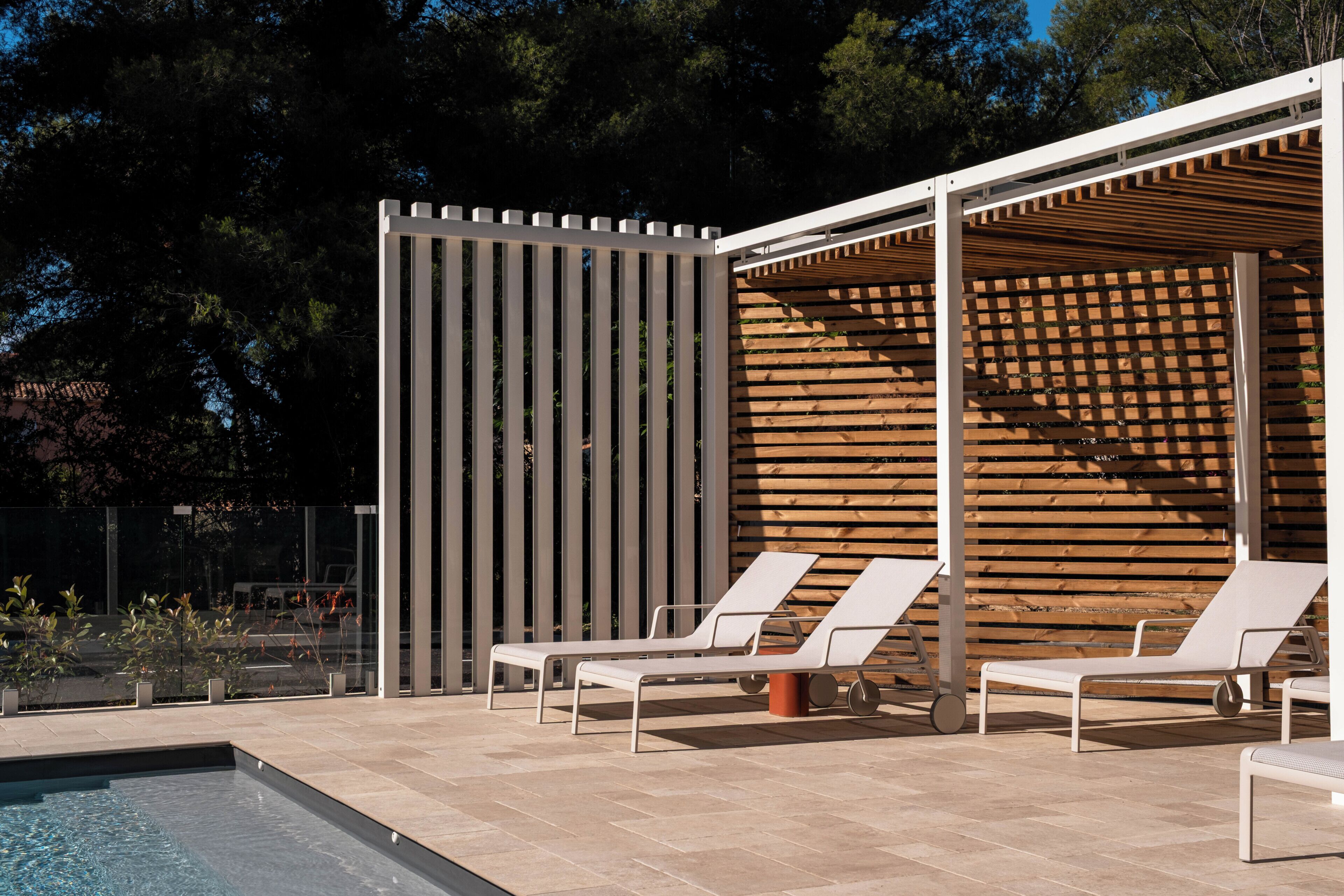 A poolside area with lounge chairs, a wooden pergola, and a glass fence.