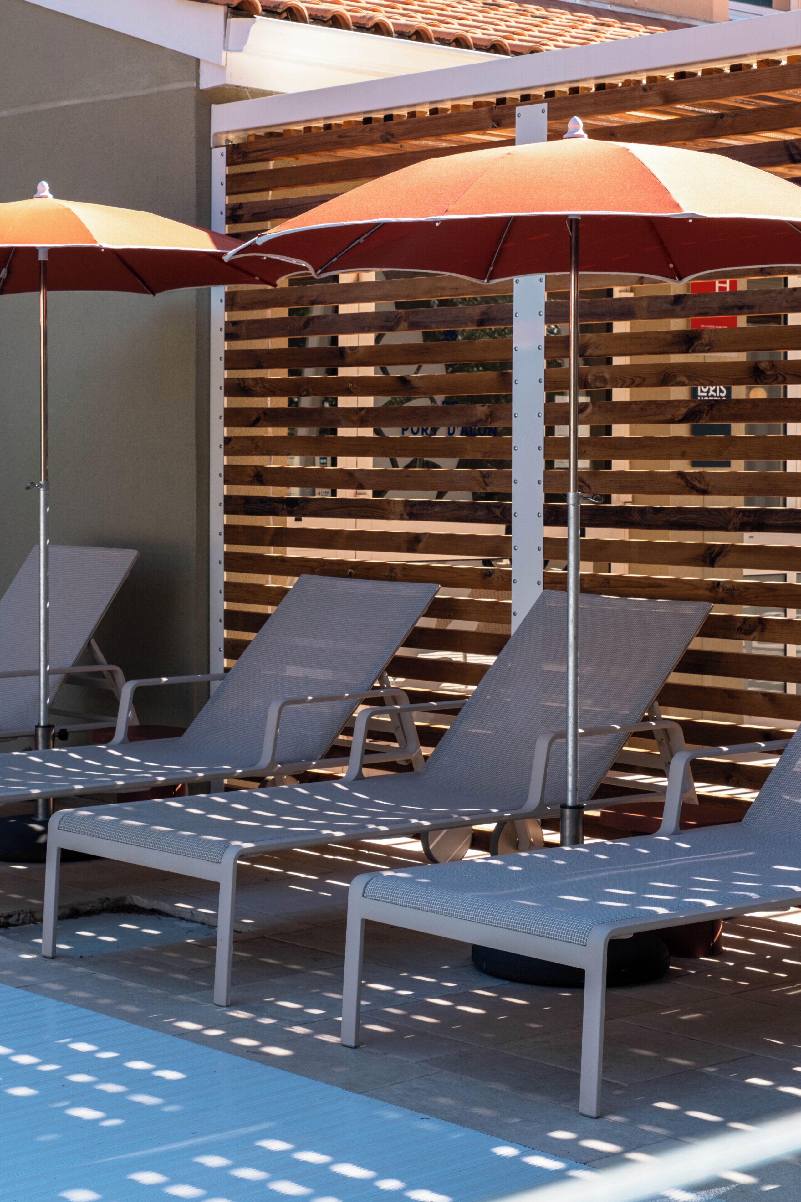 A row of sun loungers with umbrellas, a wooden slatted wall, and a metal bench.
