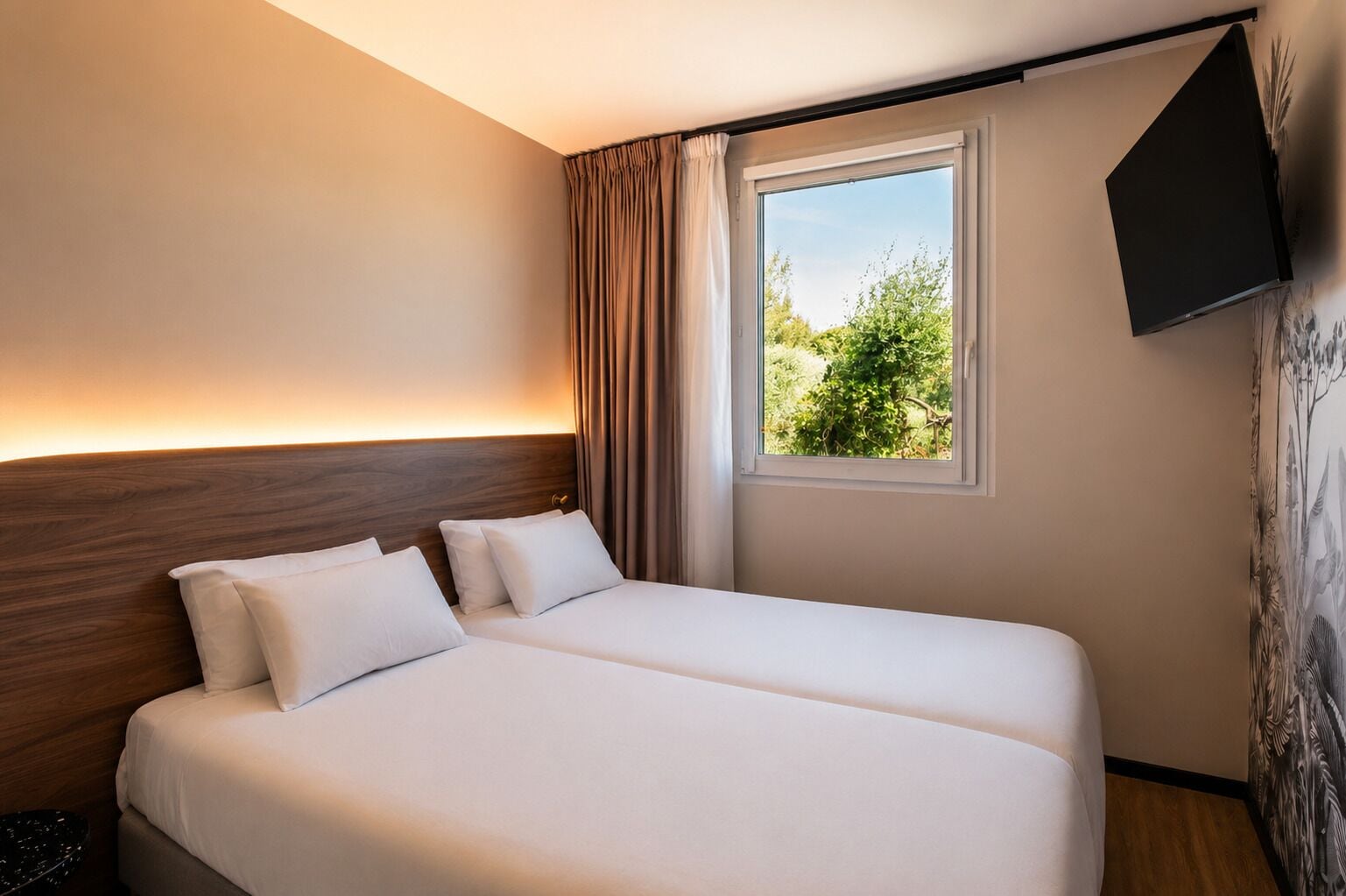 A hotel room with two beds, a window with curtains, a mounted TV, and a wall with a nature-themed mural.