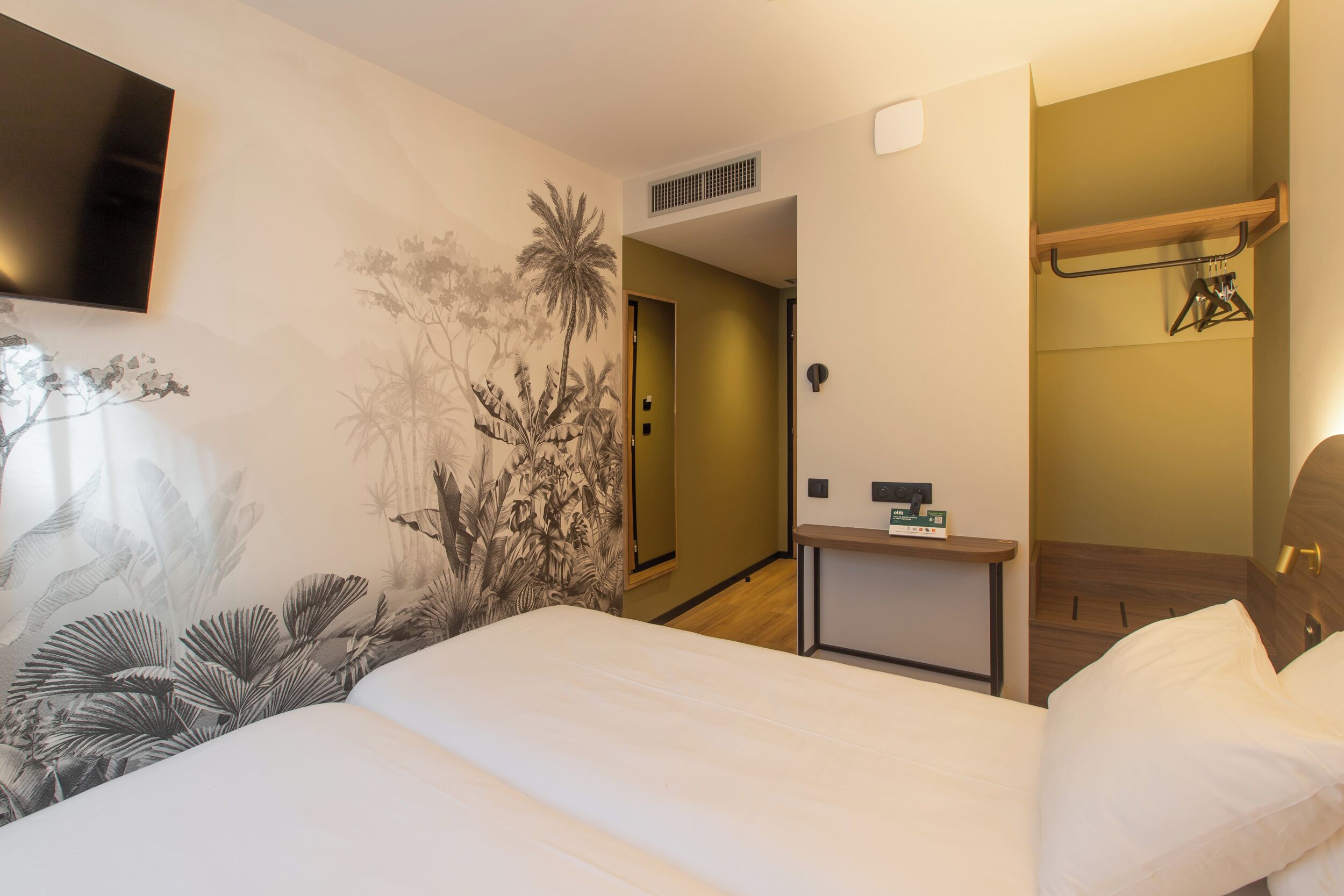 Habitación de hotel con cama, televisión montada en la pared, una mesita con un reloj y una puerta que da a otra habitación.