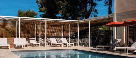 A swimming pool with lounge chairs and umbrellas, surrounded by a wooden pergola and trees.