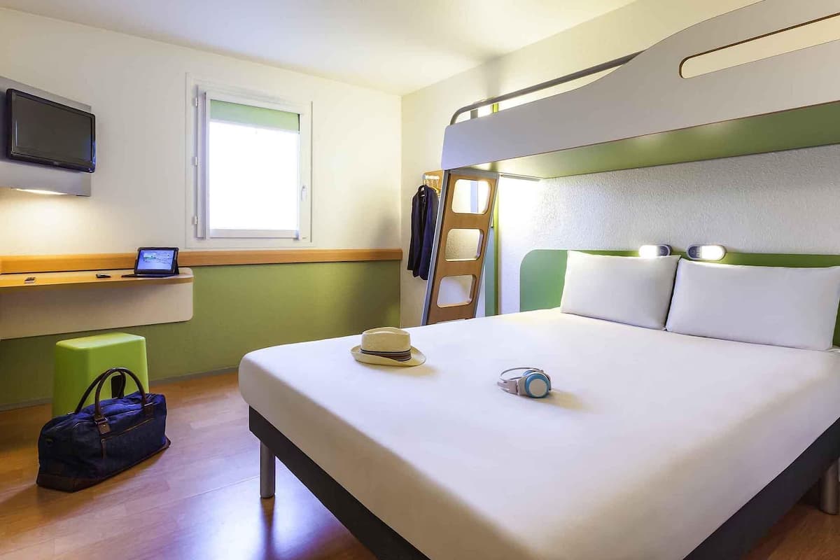 A hotel room with a bunk bed, a desk with a computer, a window, a suitcase, and a bed with pillows and headphones.
