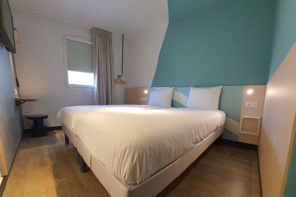 A modern hotel room with a large bed, a small bedside table, a window with curtains, and a wall-mounted light fixture.