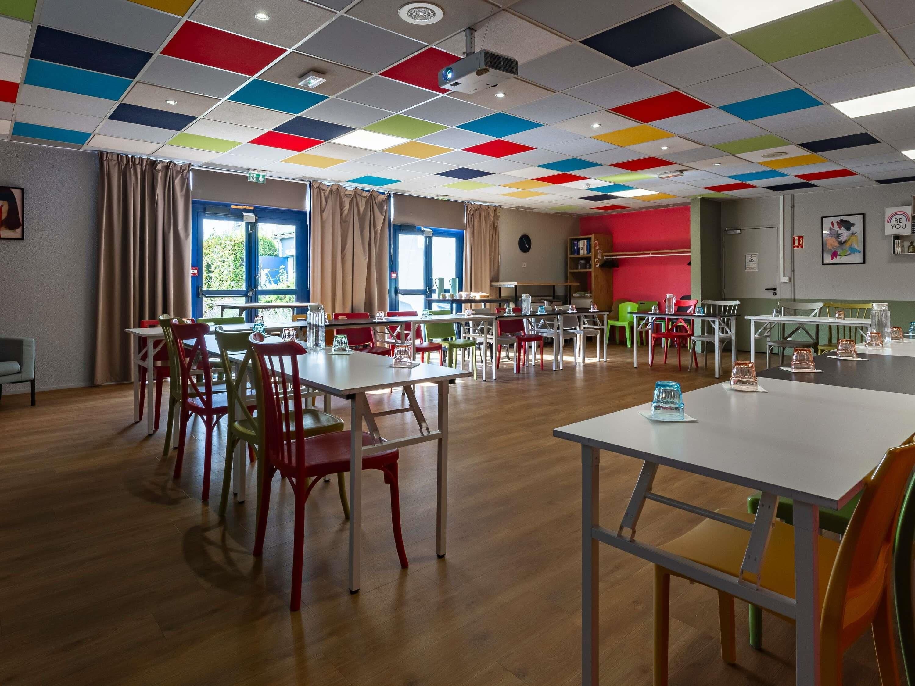 A dining area with multiple tables and chairs, a projector on the ceiling, and a red wall with a clock.