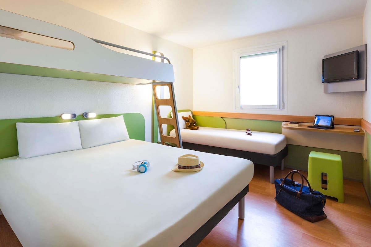 A hotel room with a bunk bed, a desk with a laptop, a TV mounted on the wall, and a window.